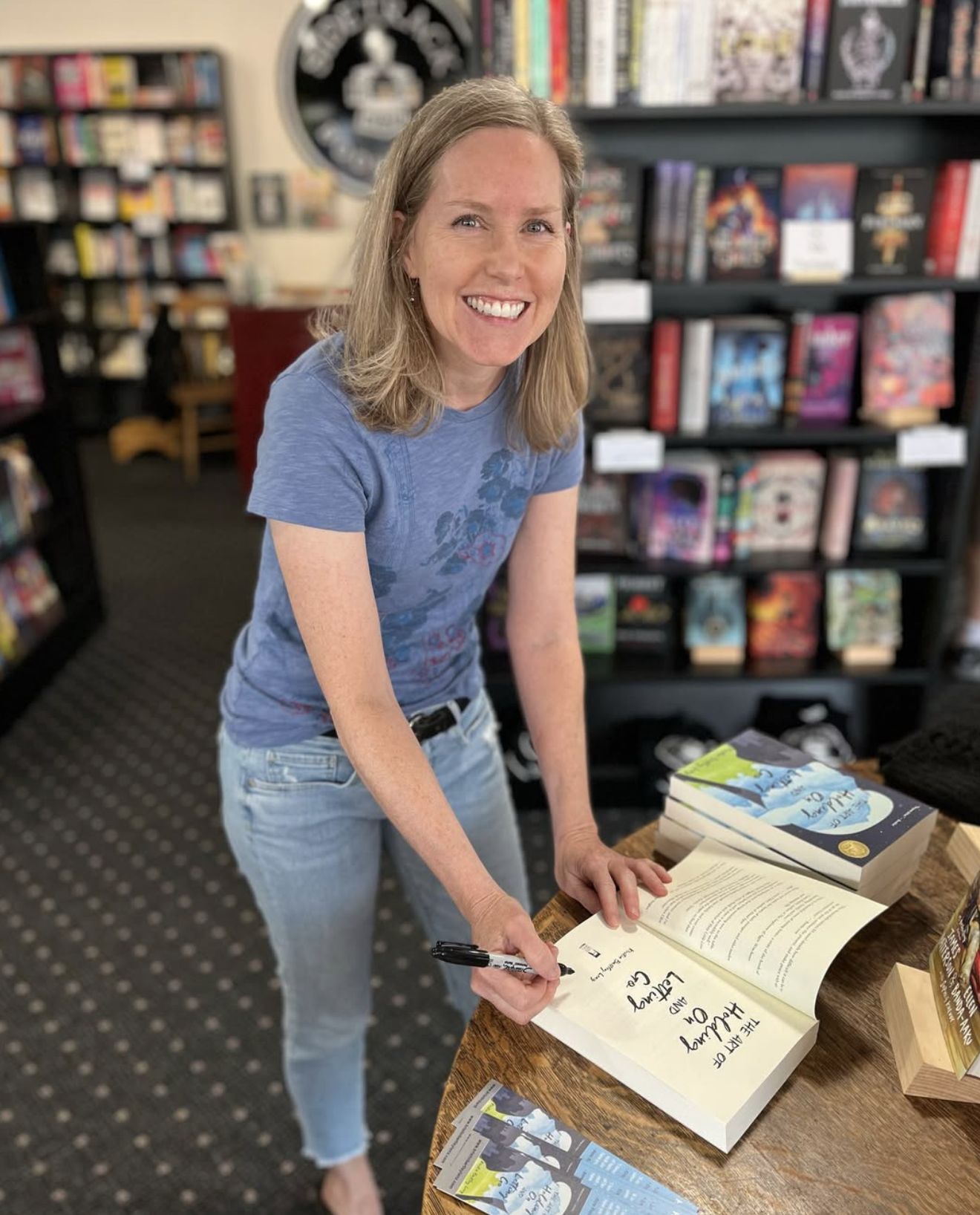 A woman with shoulder-length blonde hair, wearing a blue T-shirt and light jeans, is smiling and signing a book at a bookstore. She is standing at a wooden table filled with books and promotional materials, with shelves of books in the background.