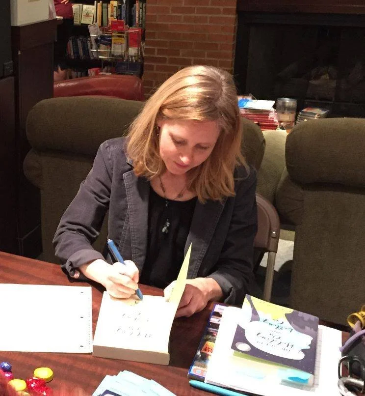A blonde woman sitting at a table, signing a book with a blue pen.