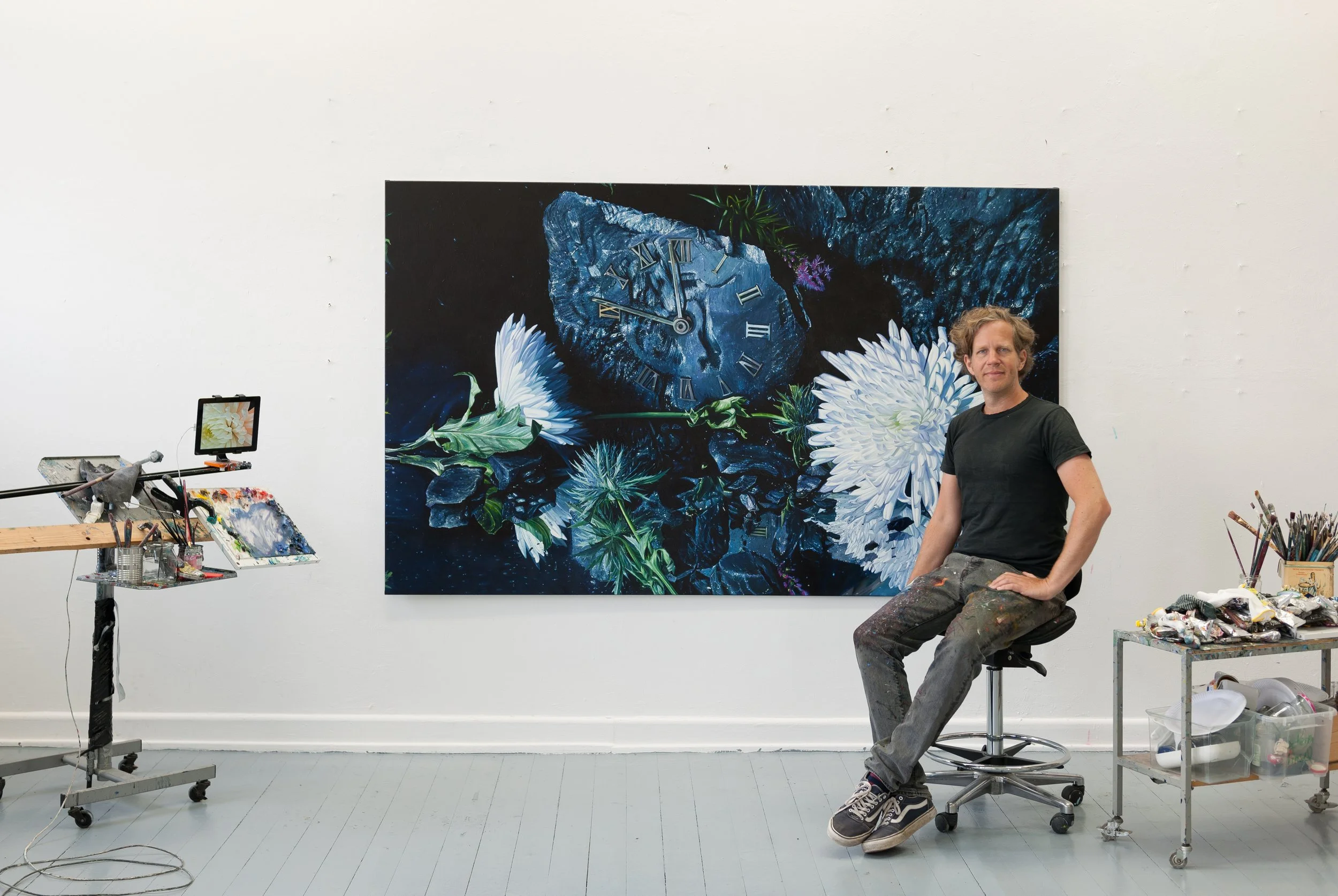 Ein Künstler sitzt vor einem großen Gemälde mit Uhren, Blumen und dunklen Naturmotiven. Er trägt eine schwarze T-Shirt und abgenutzte Hosen, umgeben von Künstlermaterialien und einem Malpult.
