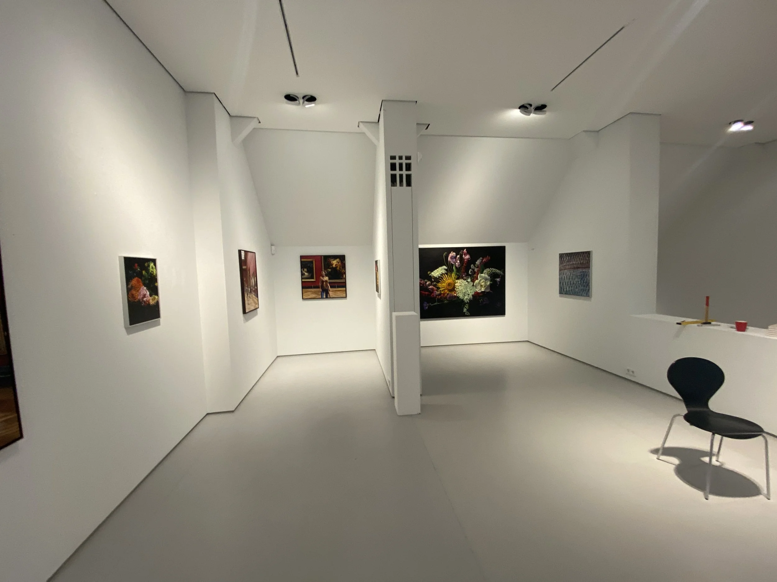 Ein weißer Galerieraum mit gedämpftem Licht, Wandkunst und einem Blumenarrangement. Es gibt schwarze Stühle und eine große Wandspiegelung.
