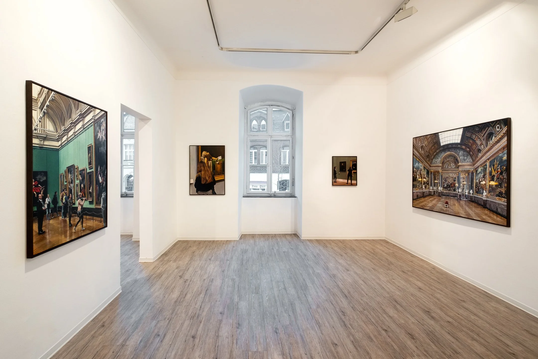 Ein Kunstraum mit weißen Wänden und Holzboden, an denen große gerahmte Gemälde hängen, darunter Szenen mit historischen oder religiösen Motiven, im Hintergrund ein Fenster mit Blick auf ein altes Gebäude.