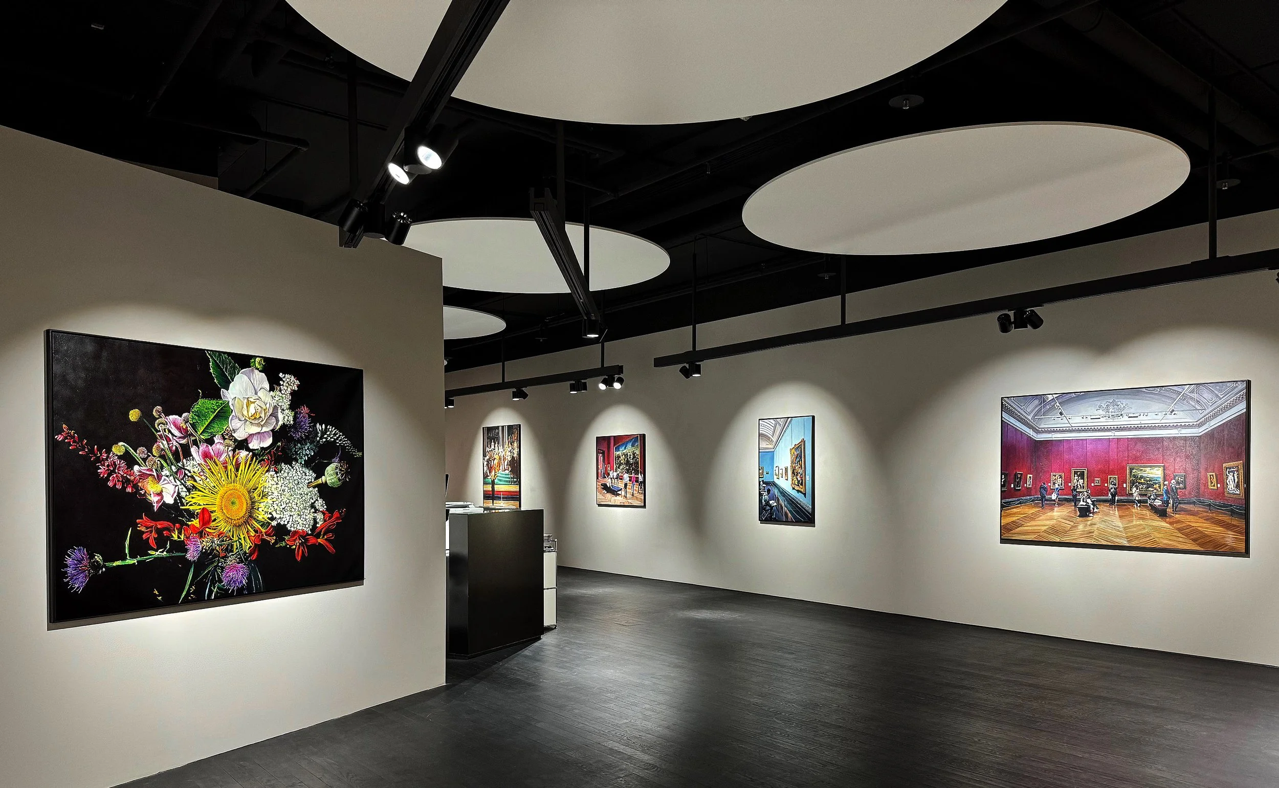 Eine Kunstgalerie mit verschiedenen gerahmten Gemälden an weißen Wänden, dunklen Deckenstrukturen und Holzboden.