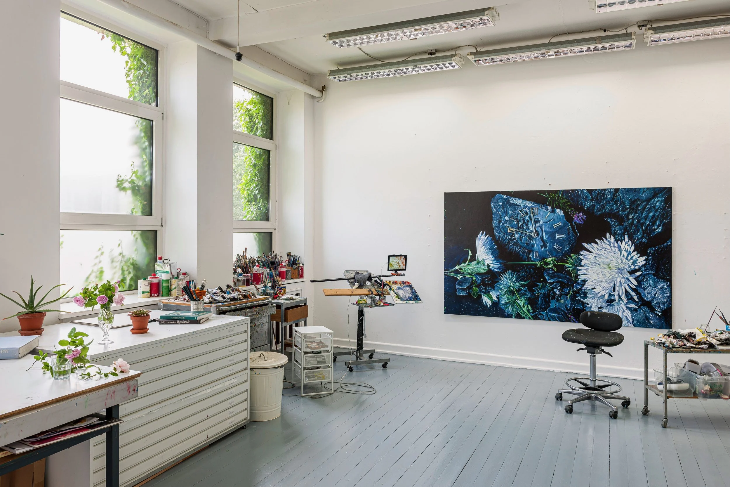 Künstleratelier mit großen Fenstern, einem großen Gemälde mit Blumen auf der Wand, Tisch mit Malmaterialien, Pflanzen und Büchern, Stuhl, Arbeitsplatz mit Stifte und Farben, viel Tageslicht.