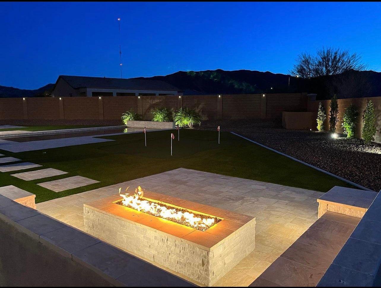Backyard at night with a rectangular fire feature on a stone patio, a well-maintained lawn with putting holes marked by flags, and landscaped areas with plants and trees, illuminated by outdoor lighting.