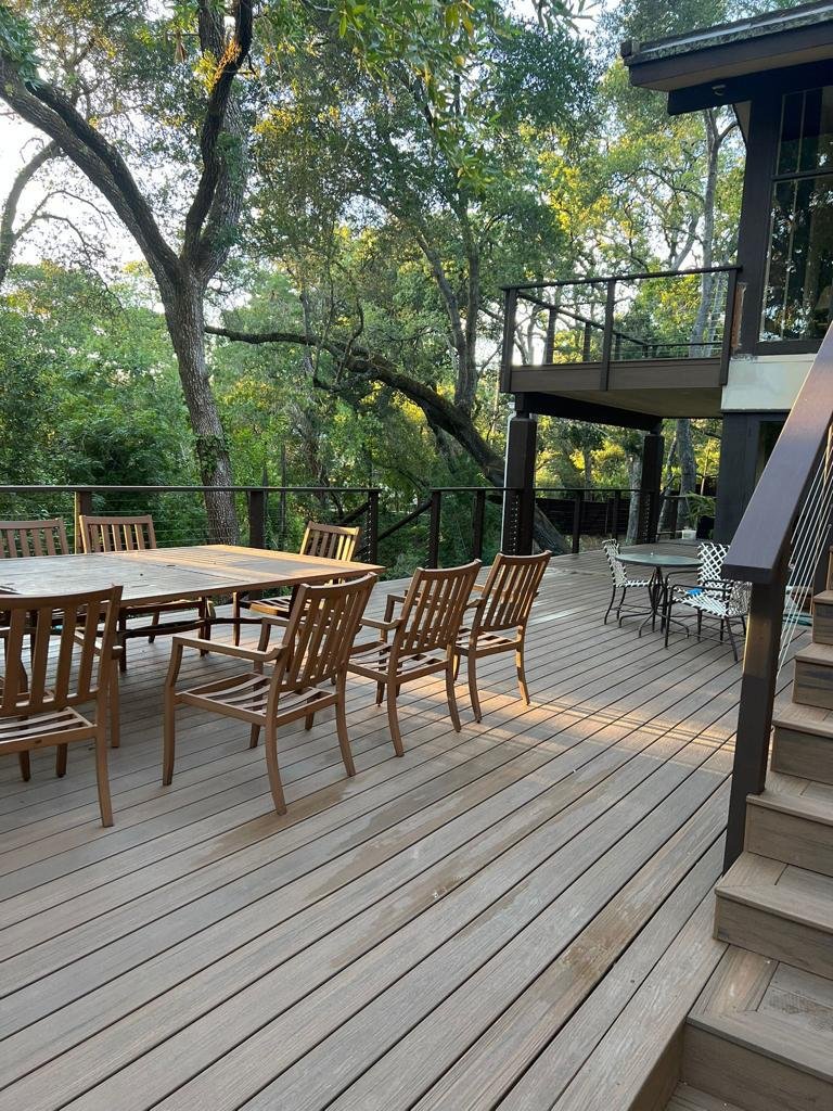 Wooden deck with outdoor dining furniture surrounded by trees, with stairs leading up to a higher level.