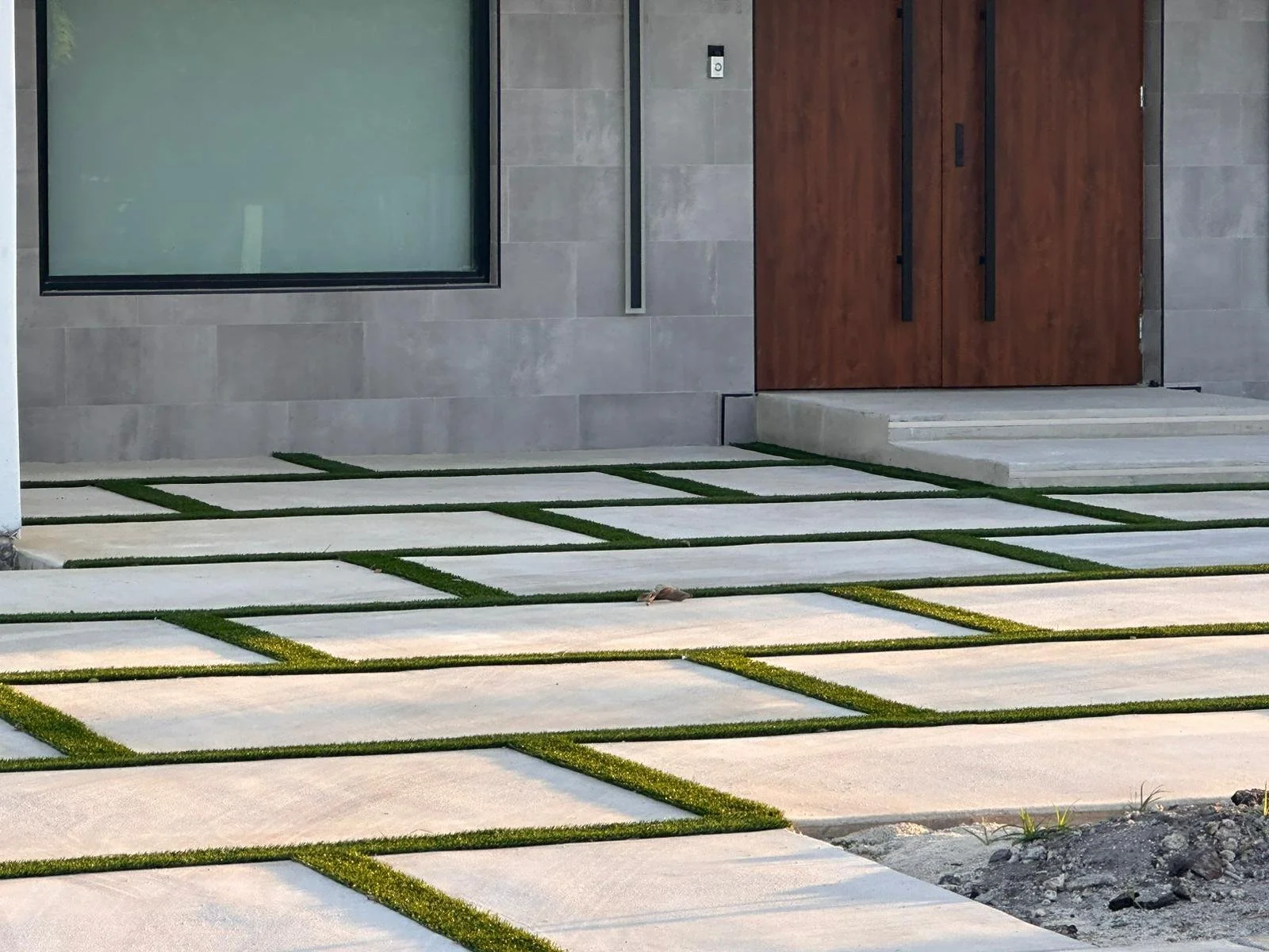 Modern concrete front walkway with grass between the concrete slabs, leading to a wooden double door entrance of a house, with a large window on the left side.
