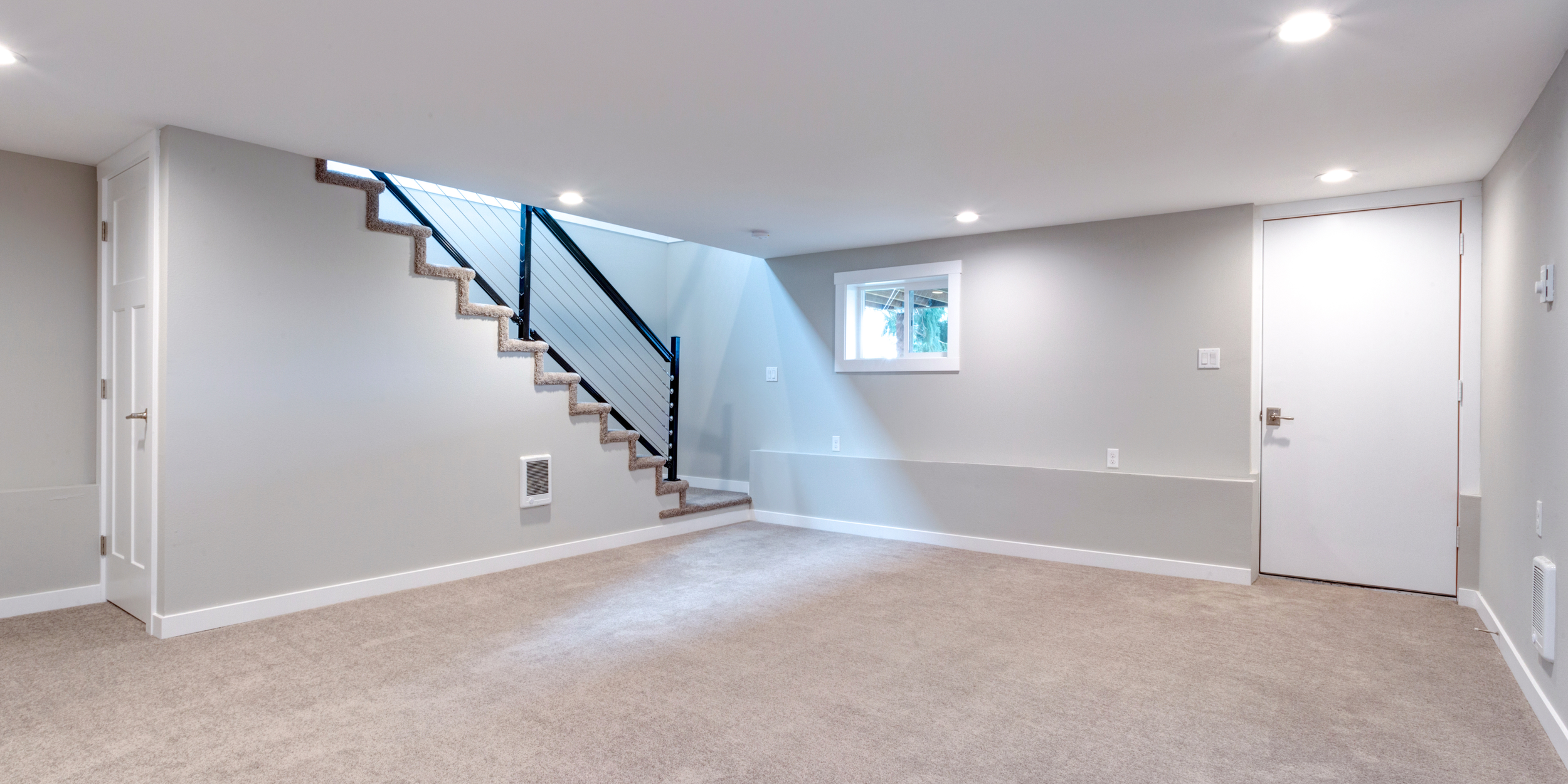 Empty basement room with beige carpet, white walls, small window, staircase with black railing, door on left, ceiling lights.