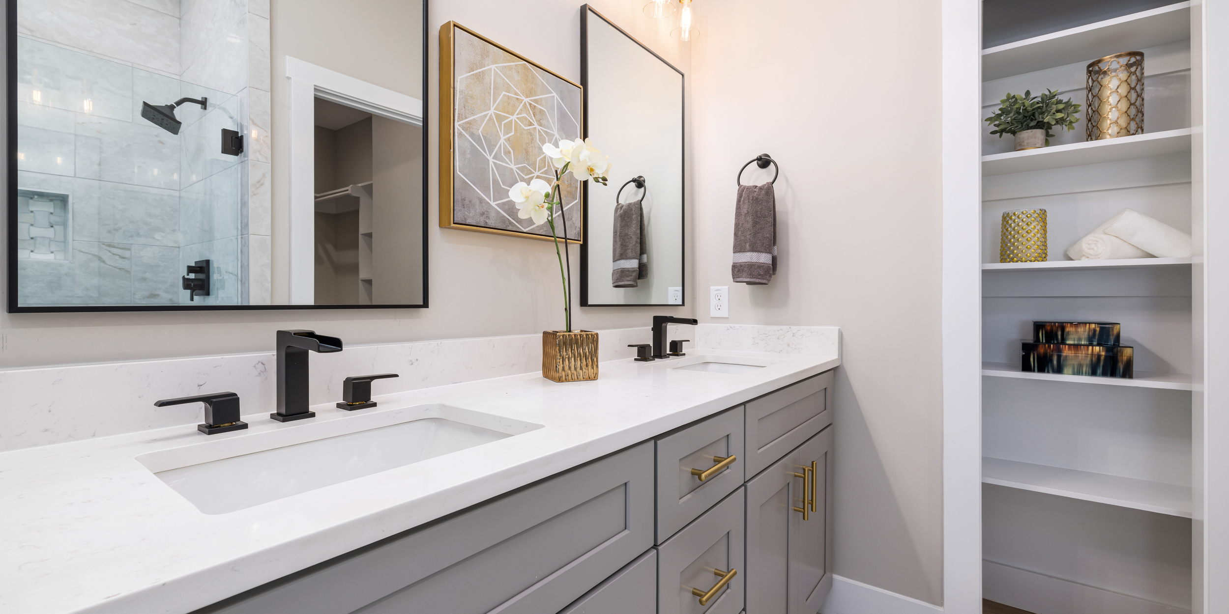Modern bathroom vanity with two sinks, black fixtures, and a gray Cabinet with gold hardware. A large mirror with a black frame, a decorative orchid in a gold vase, and two framed abstract art pieces hang on the wall. A towel ring with a gray towel is next to each mirror. To the right, white open shelving with decorative items including plants, rolled towels, and a black and gold box.