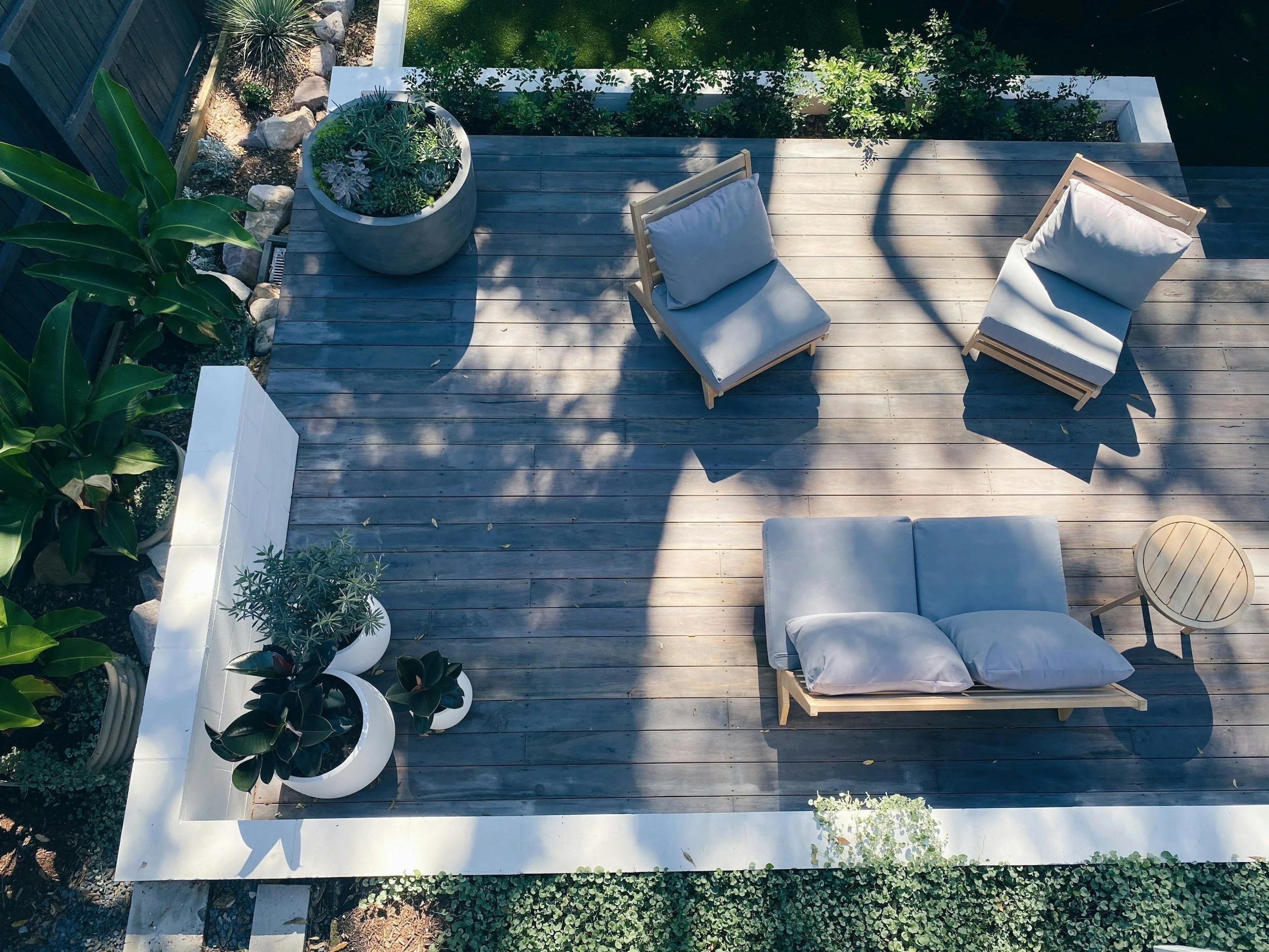 An outdoor deck with plants and seating furniture, including a small round table, a cushioned lounge chair, two cushioned chairs, and a white border surrounding the deck.