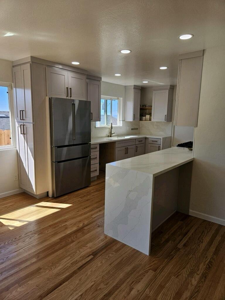 Modern kitchen with white and gray cabinets, stainless steel refrigerator, wooden floor, white marble countertop, and natural light coming through windows.