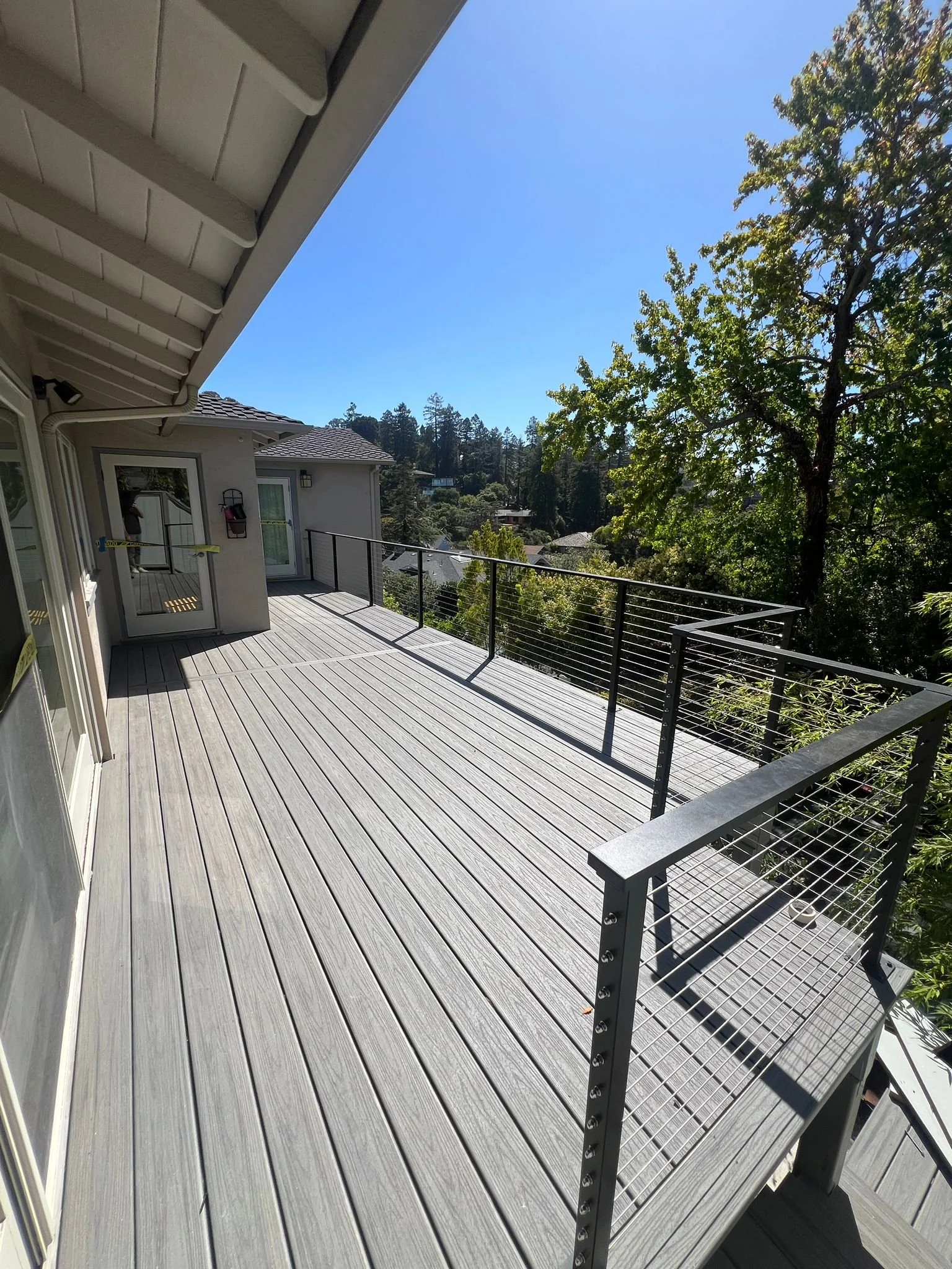Empty outdoor wooden deck with black metal railing, attached to a house, with trees and houses in the background under a clear blue sky.