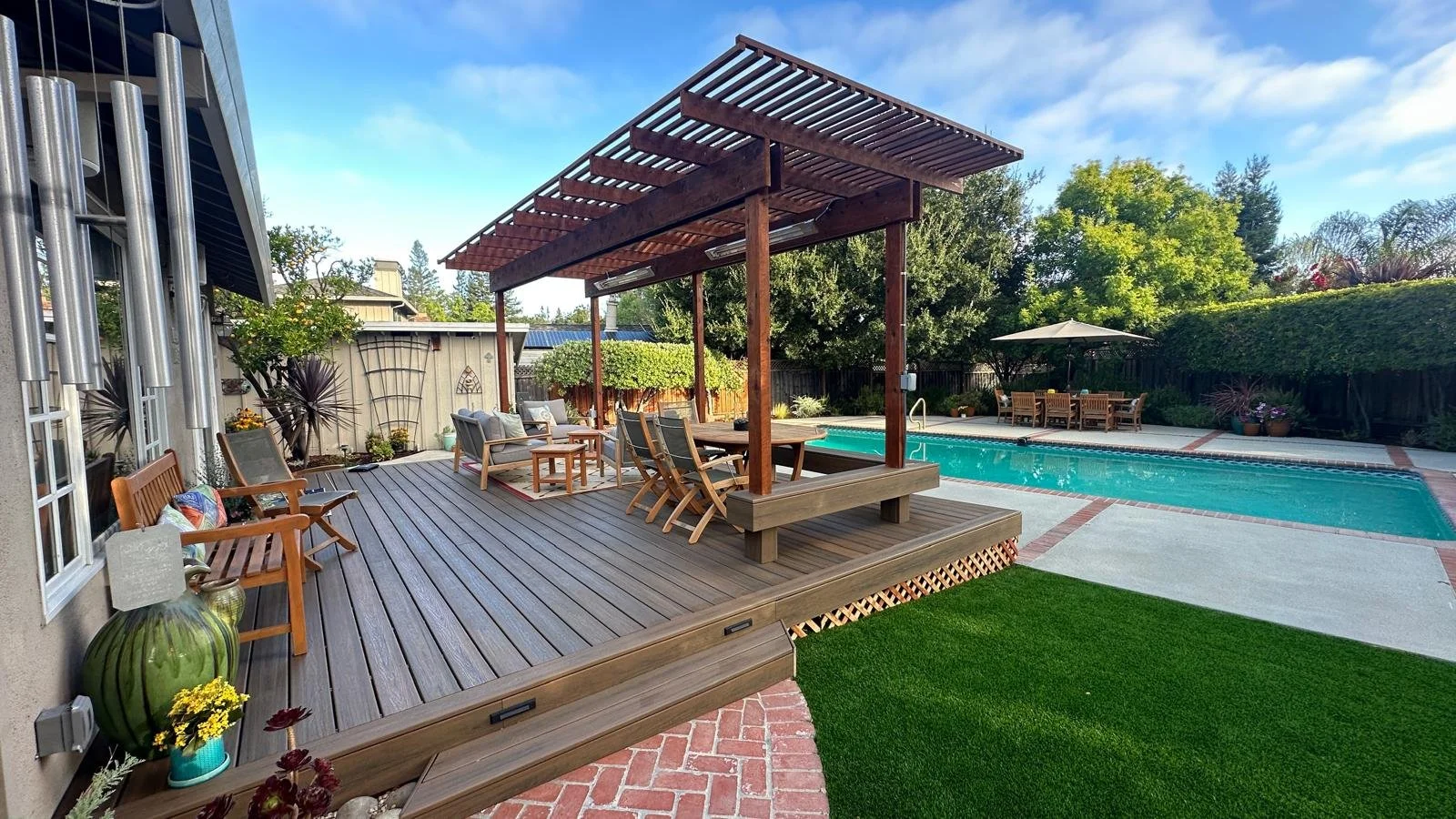 Backyard with swimming pool, wooden deck, pergola, outdoor furniture, and lush greenery.