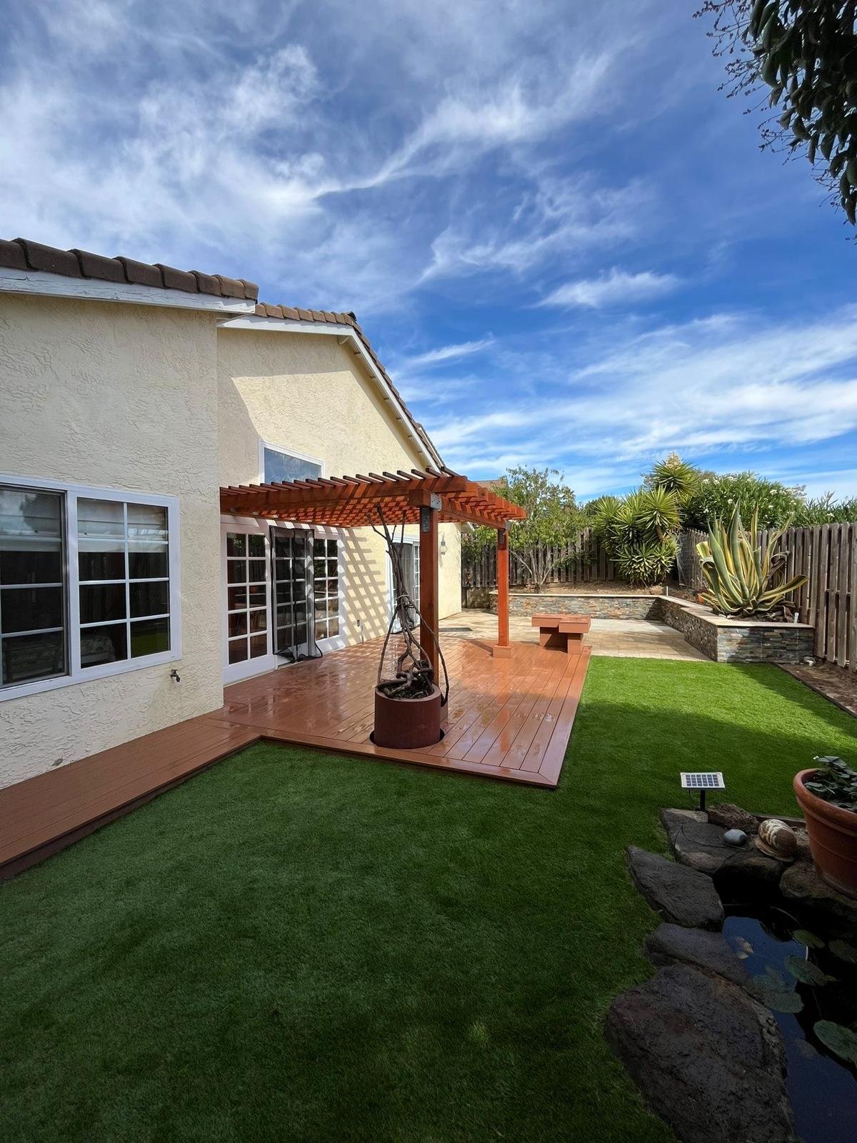 A backyard with a wooden deck, artificial grass, a small pond with rocks, potted plants, and a house with white-framed windows. The sky is blue with wispy clouds.
