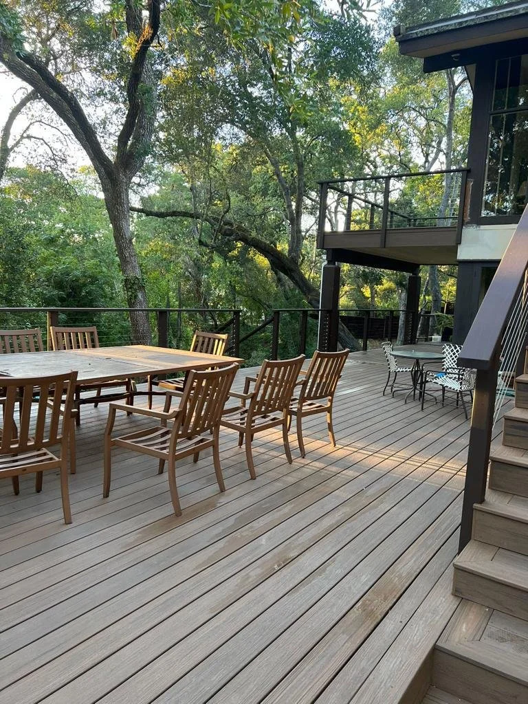 Wooden deck with outdoor dining table and chairs, surrounded by trees and greenery, part of a modern house with stairs and upper balcony.