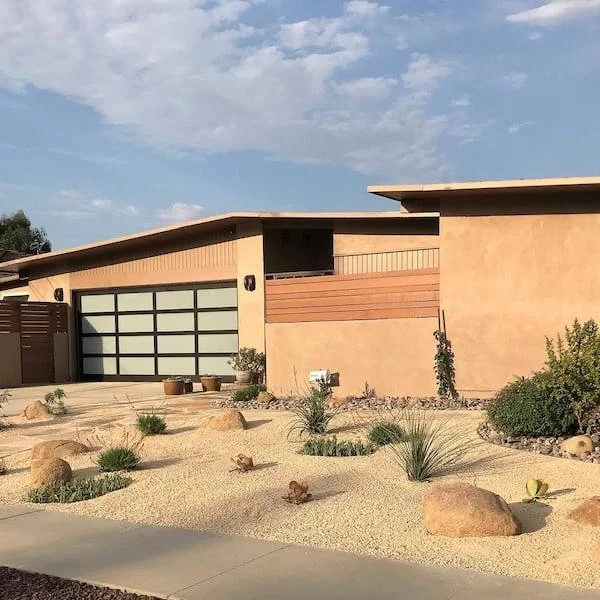 Modern house with desert landscaping including rocks, succulents, and sparse plants under a blue sky.