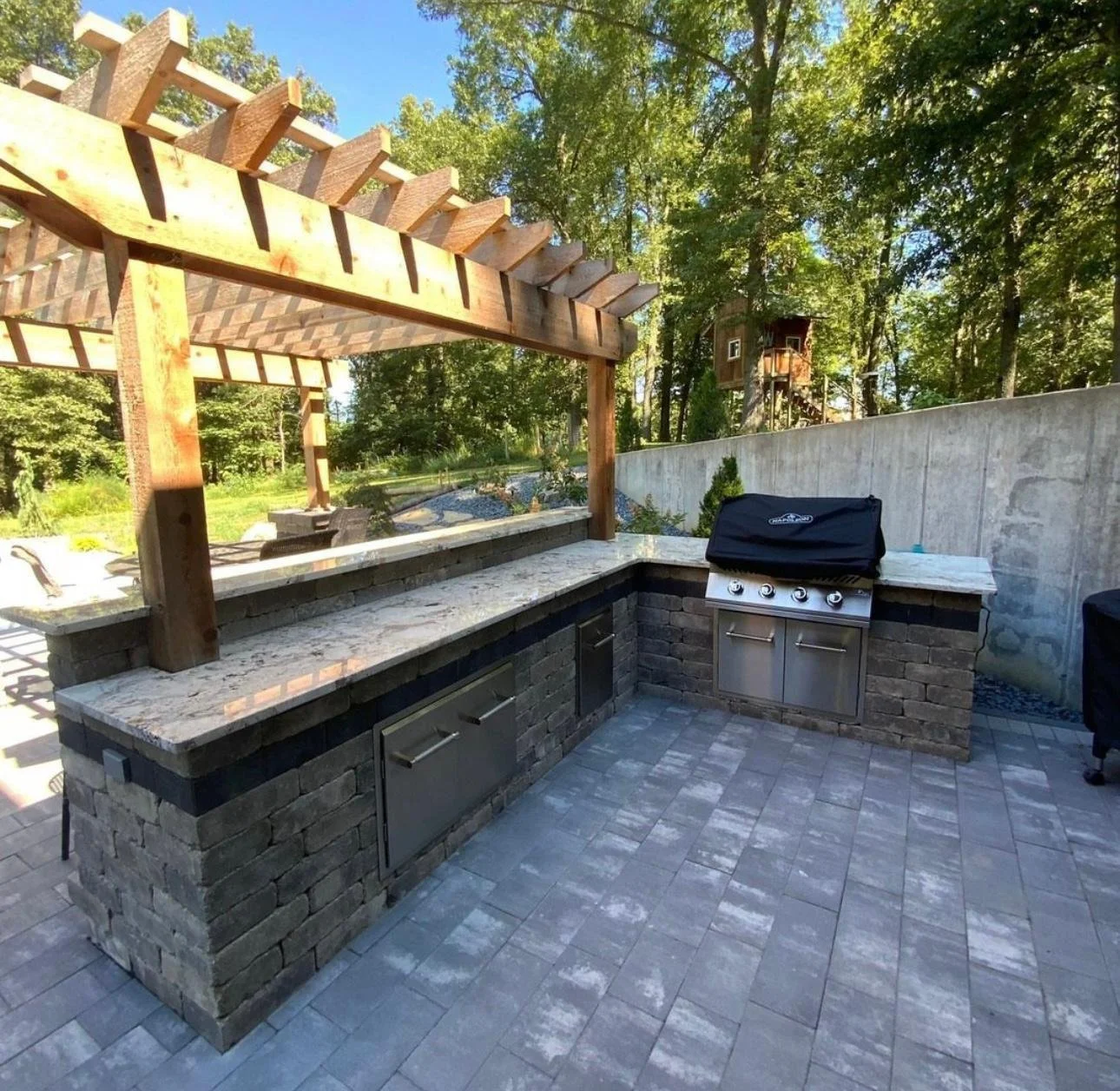 Outdoor kitchen with a built-in grill and countertop, surrounded by trees and a wooden pergola overhead.