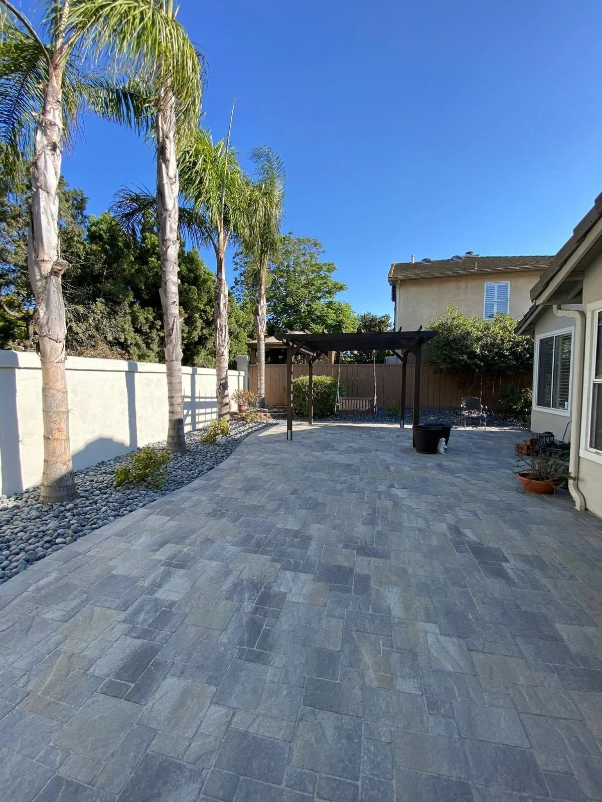 A backyard patio with gray paver stones, several tall palm trees beside a white fence, a black pergola, some potted plants, a small lawn area, and a neighboring house under a bright blue sky.