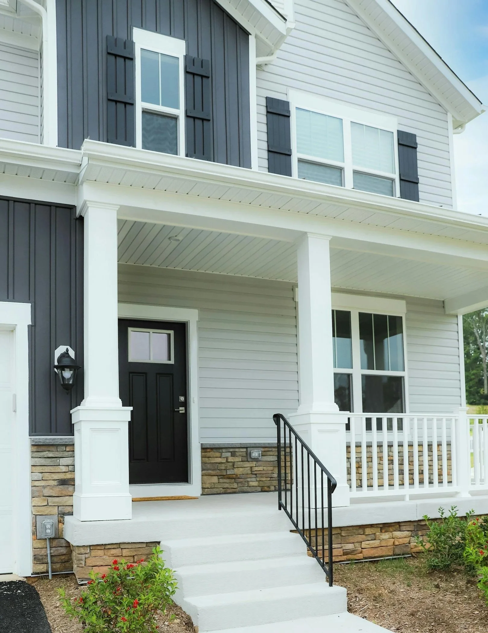 Front view of a modern two-story house with gray siding, black shutters, a black front door, a small front porch, and a staircase leading up to the porch.