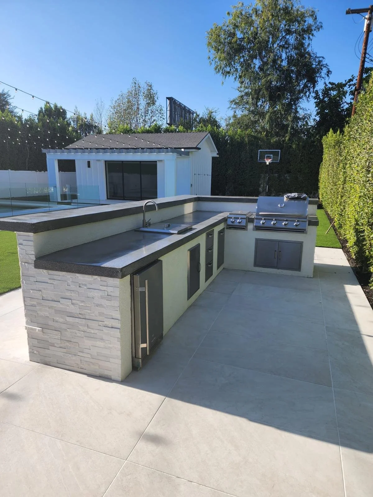 Outdoor kitchen with built-in grill, sink, and cabinets on a patio, with a small shed or guest house, basketball hoop, and trees in the background.
