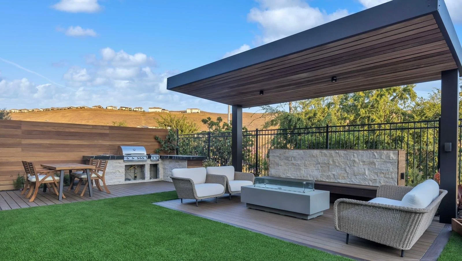 Outdoor rooftop patio with seating area, fireplace, wooden dining table, gas grill, and artificial grass, with hills and houses in the background under partly cloudy sky.