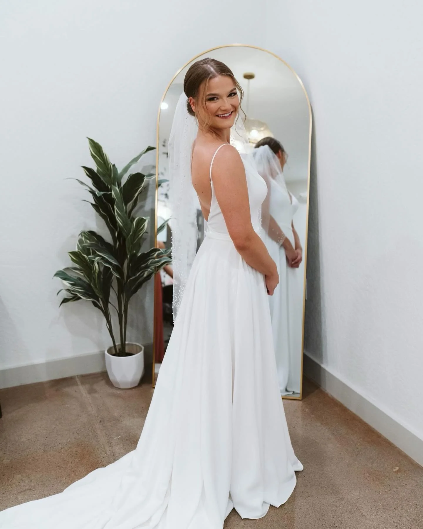 A bride in a white wedding dress with thin straps, smiling, standing in front of a mirror, near a potted plant.