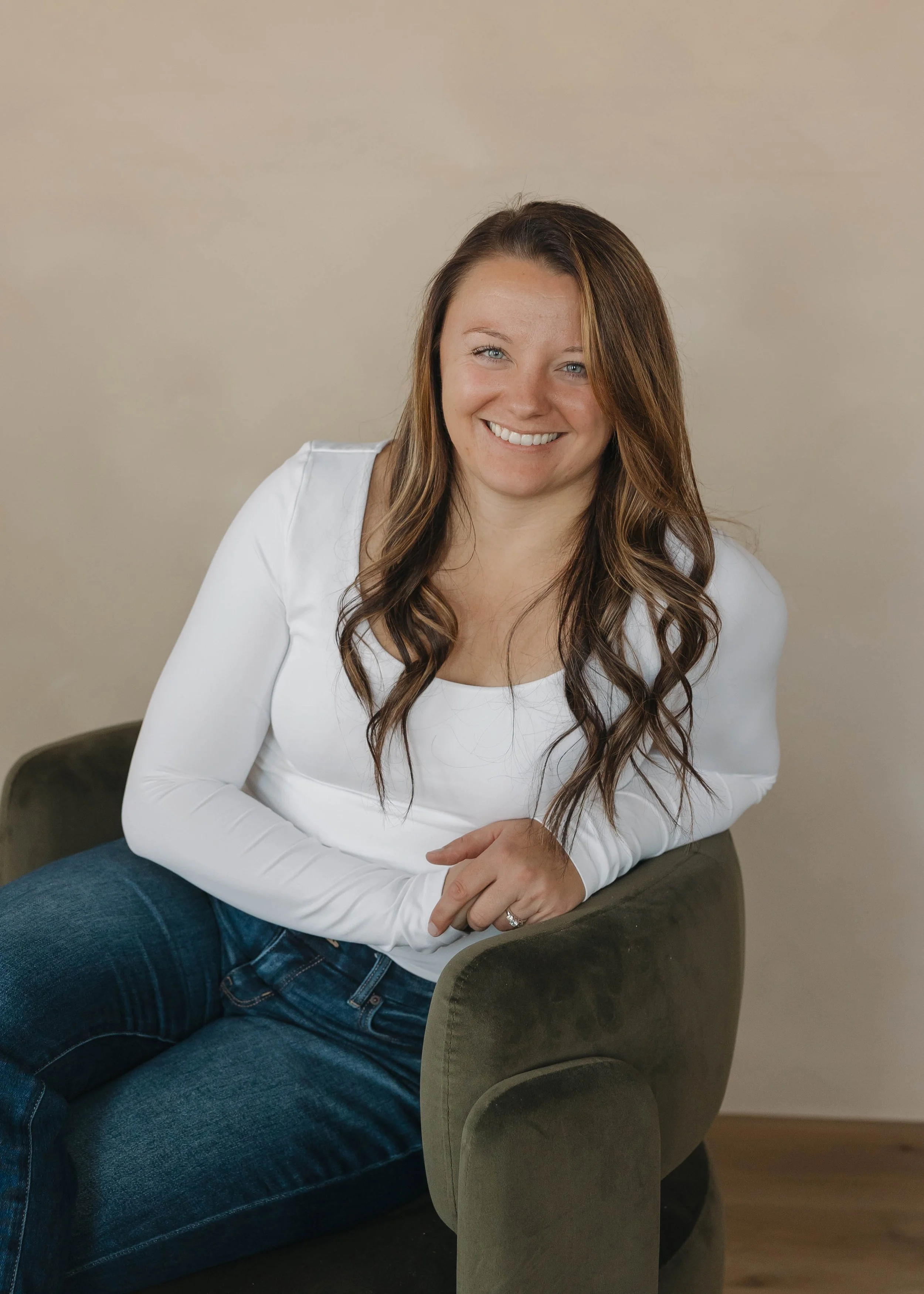 A woman with long wavy brown hair, wearing a white long-sleeve top and blue jeans, sitting on a green armchair, smiling at the camera.