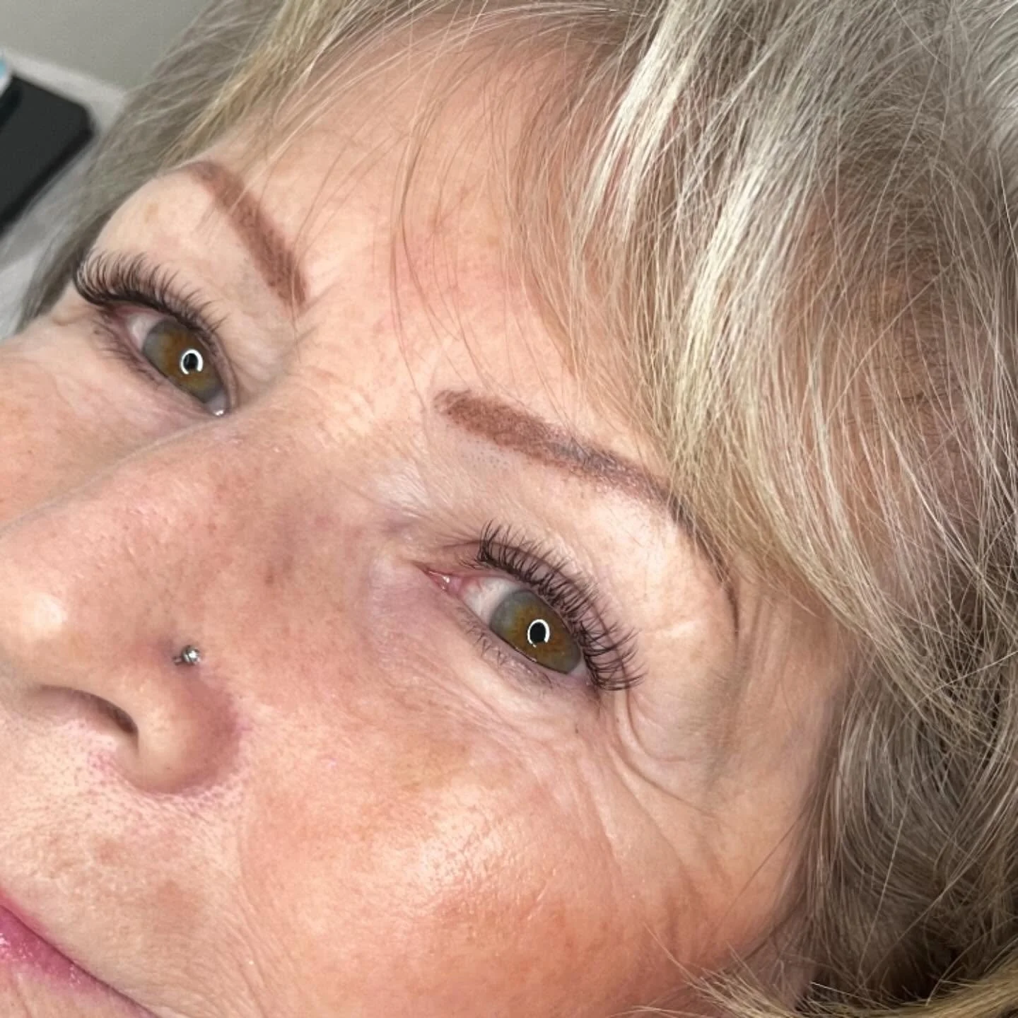 Close-up of a smiling woman with blonde hair, hazel eyes, a nose piercing, and well-groomed eyebrows.