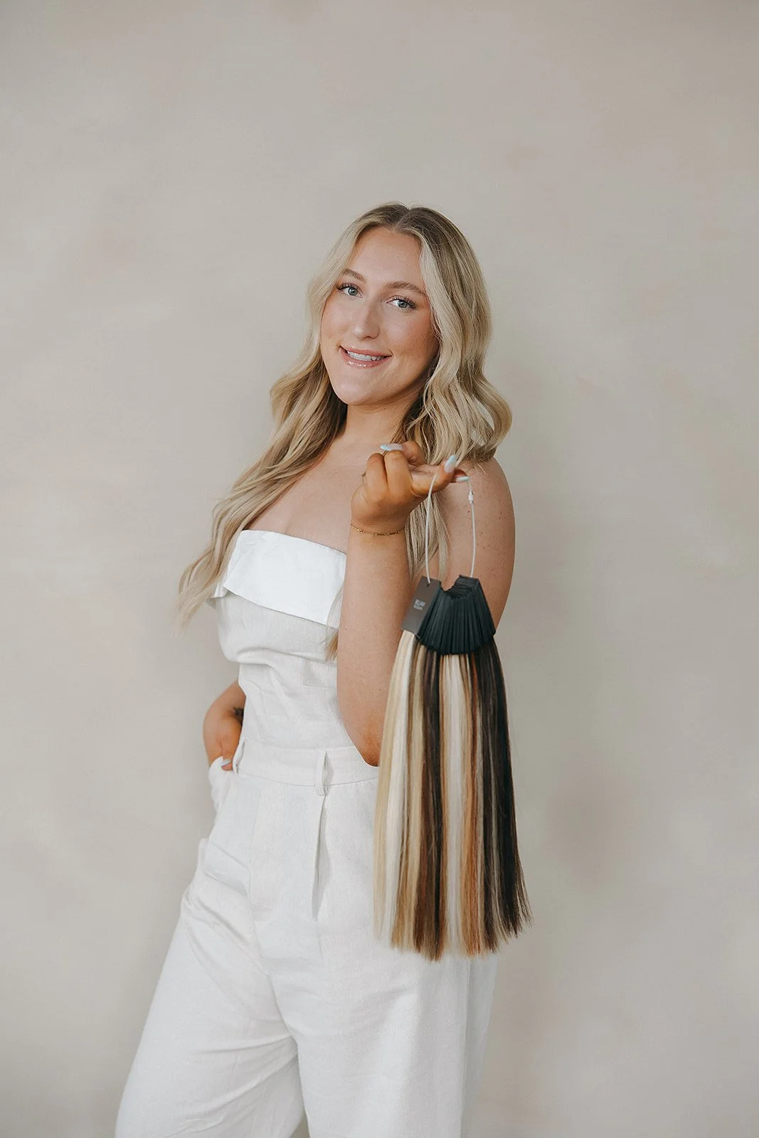 A woman with long blonde hair holding a hair color swatch fan, smiling at the camera, wearing a white strapless outfit, against a plain light-colored background.