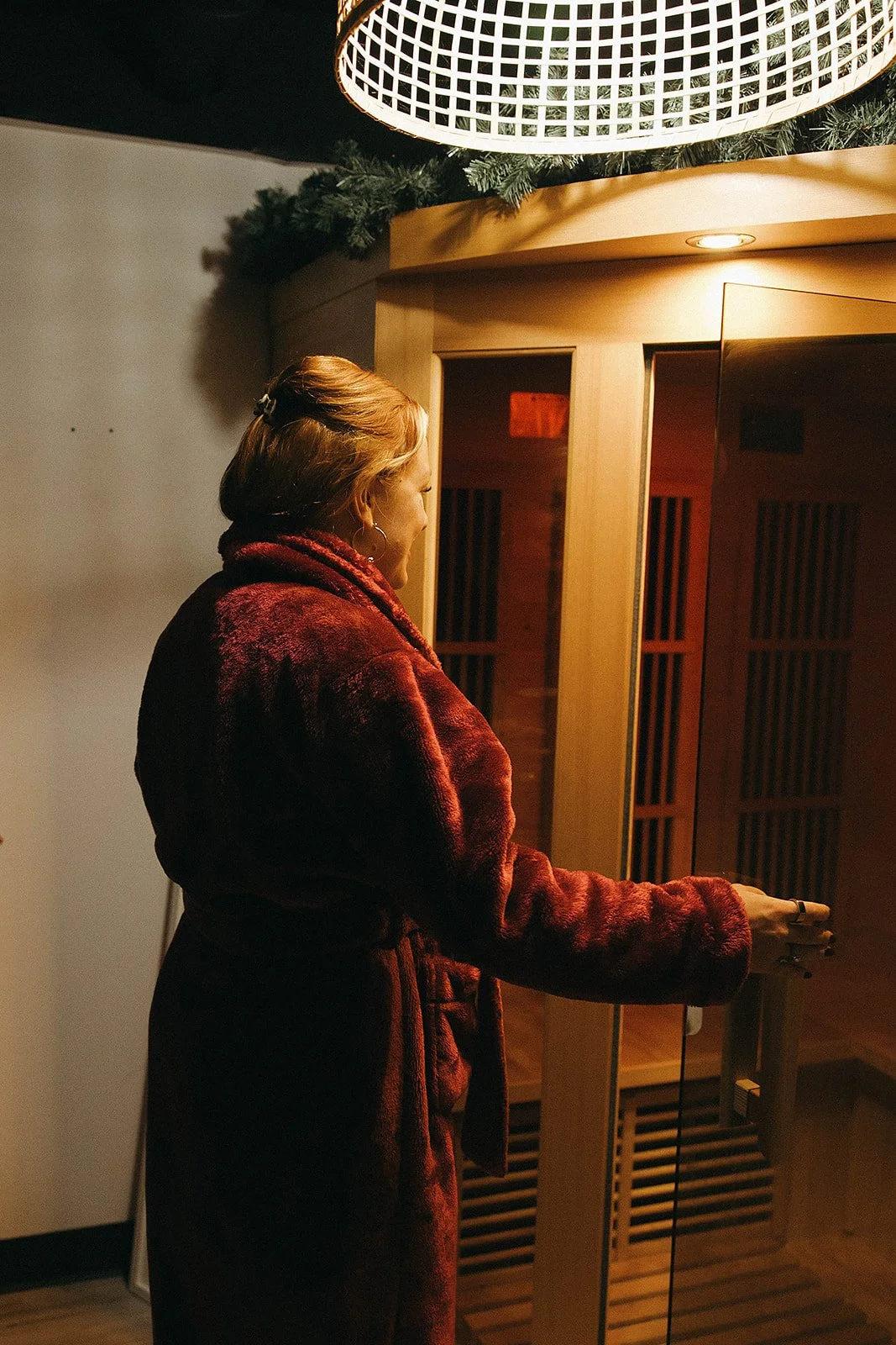 A woman in a red coat in a meditation or relaxation room, holding a small bell or chime, standing next to a wooden structure with slats and a glass panel.