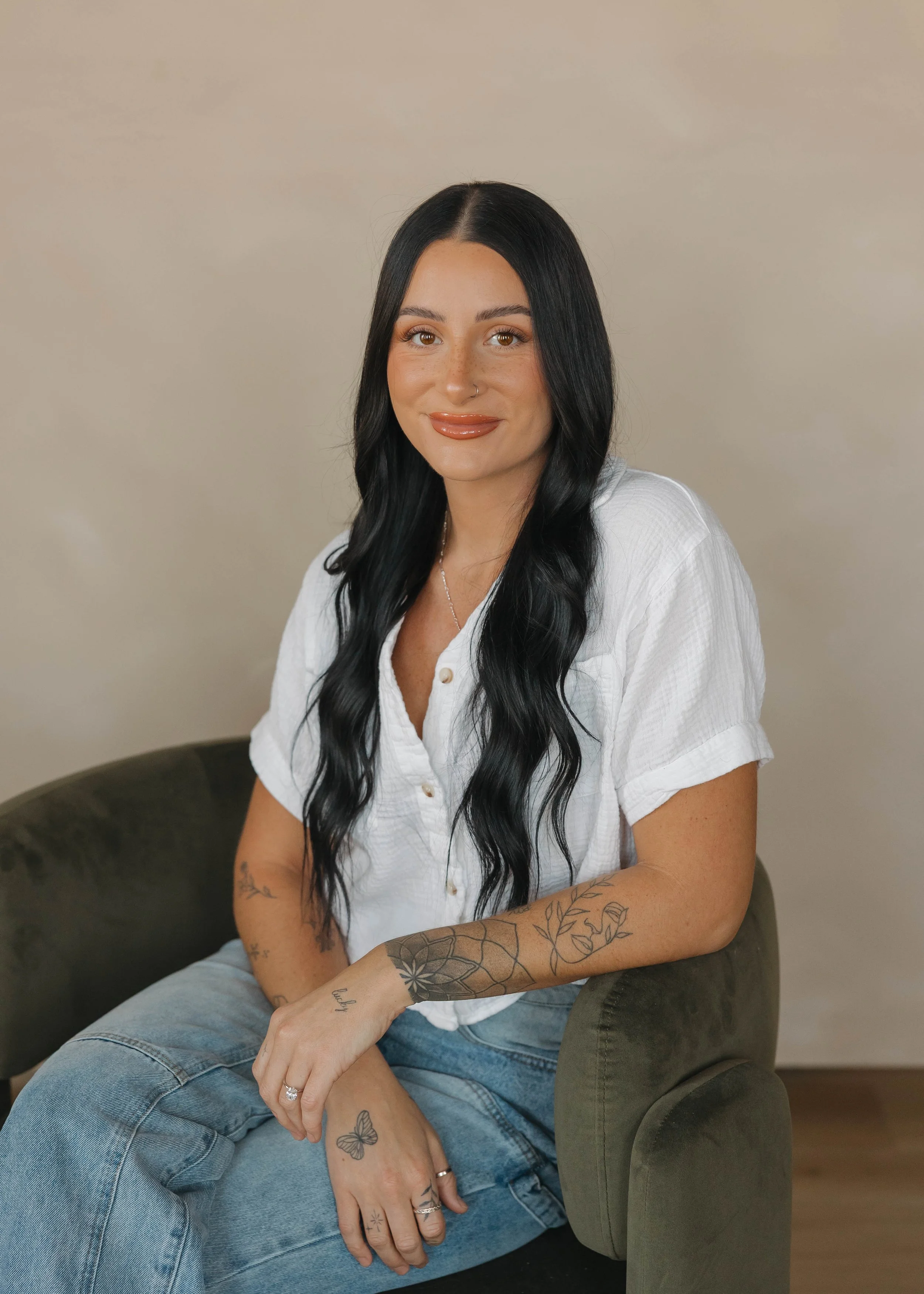 A woman with long black hair, wearing a white button-up shirt and light blue jeans, sitting on a dark green armchair against a plain wall.