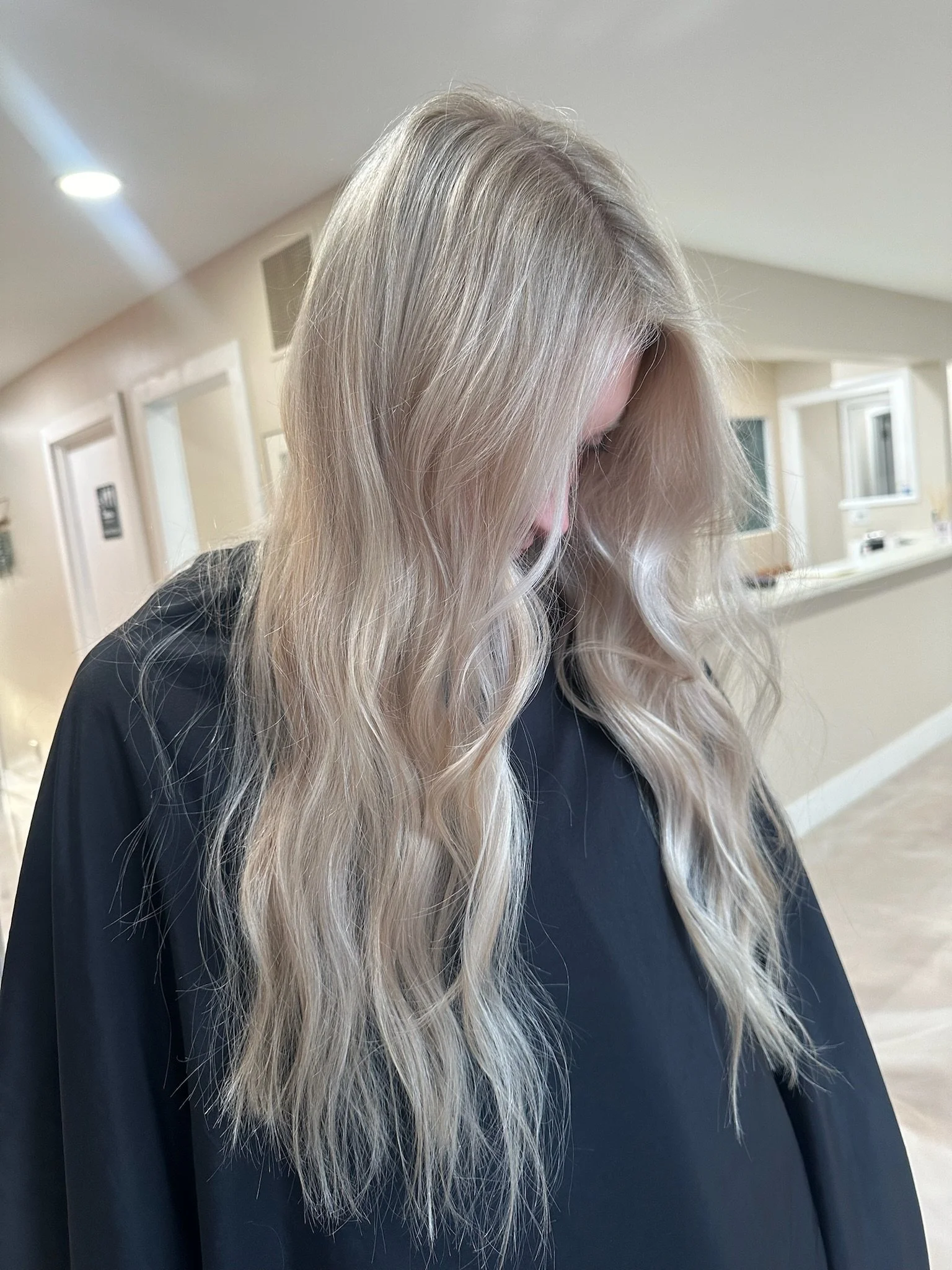 A woman with long, wavy blonde hair standing indoors, looking down, wearing a black cape.
