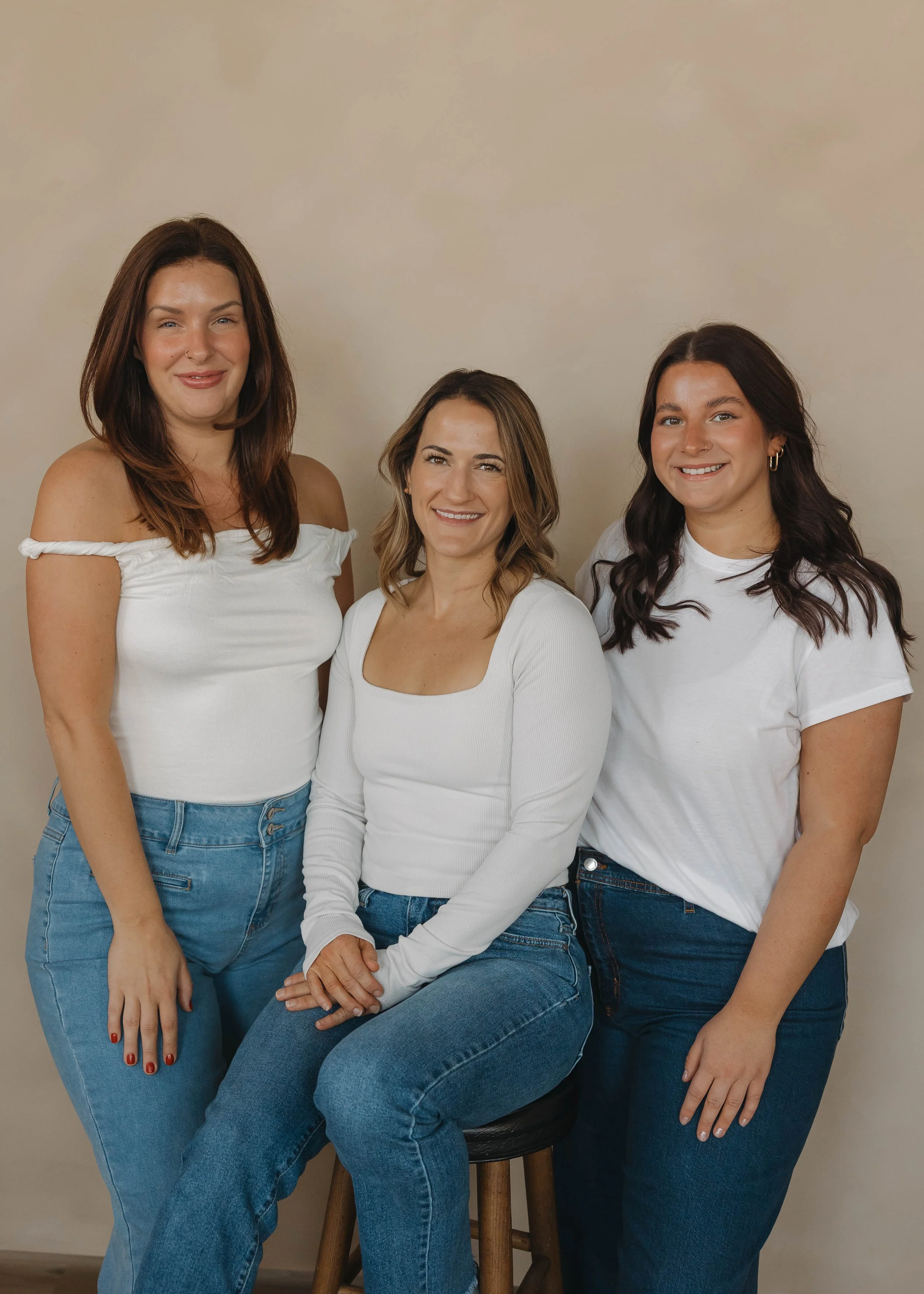 Three women in casual white tops and jeans posing together against a beige background. One woman is seated on a stool, while the other two stand beside her, smiling at the camera.