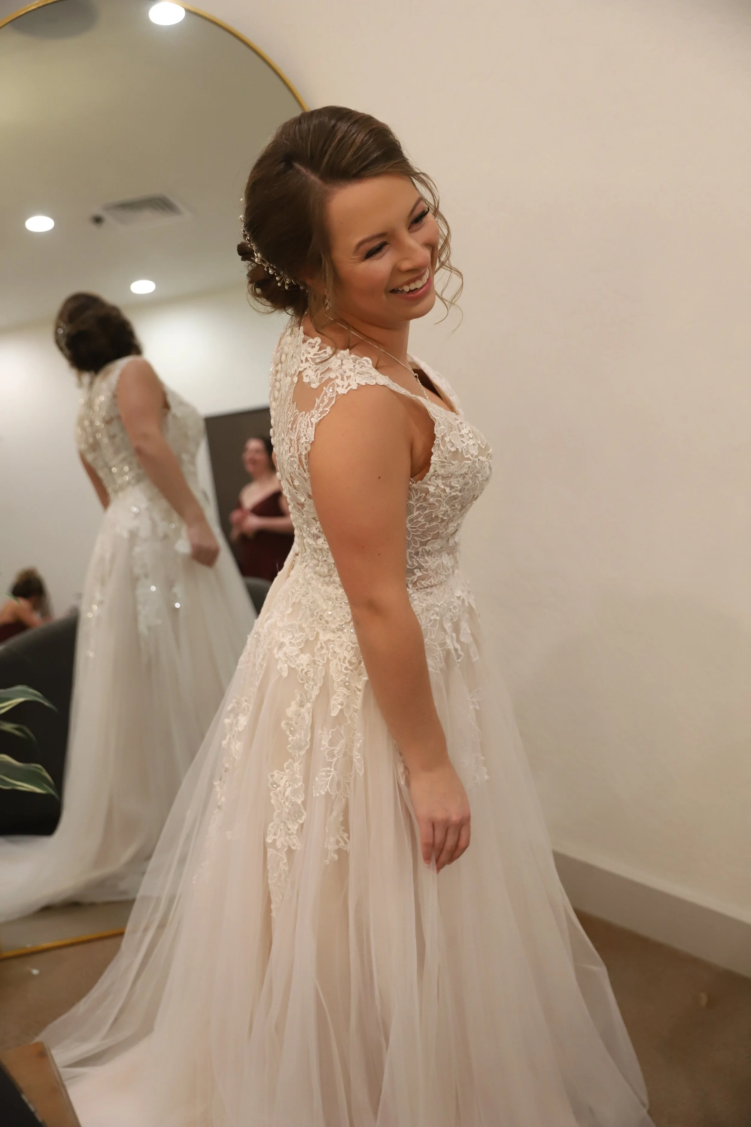 Woman trying on a wedding dress in a fitting room, smiling, with a mirror and other people in the background.