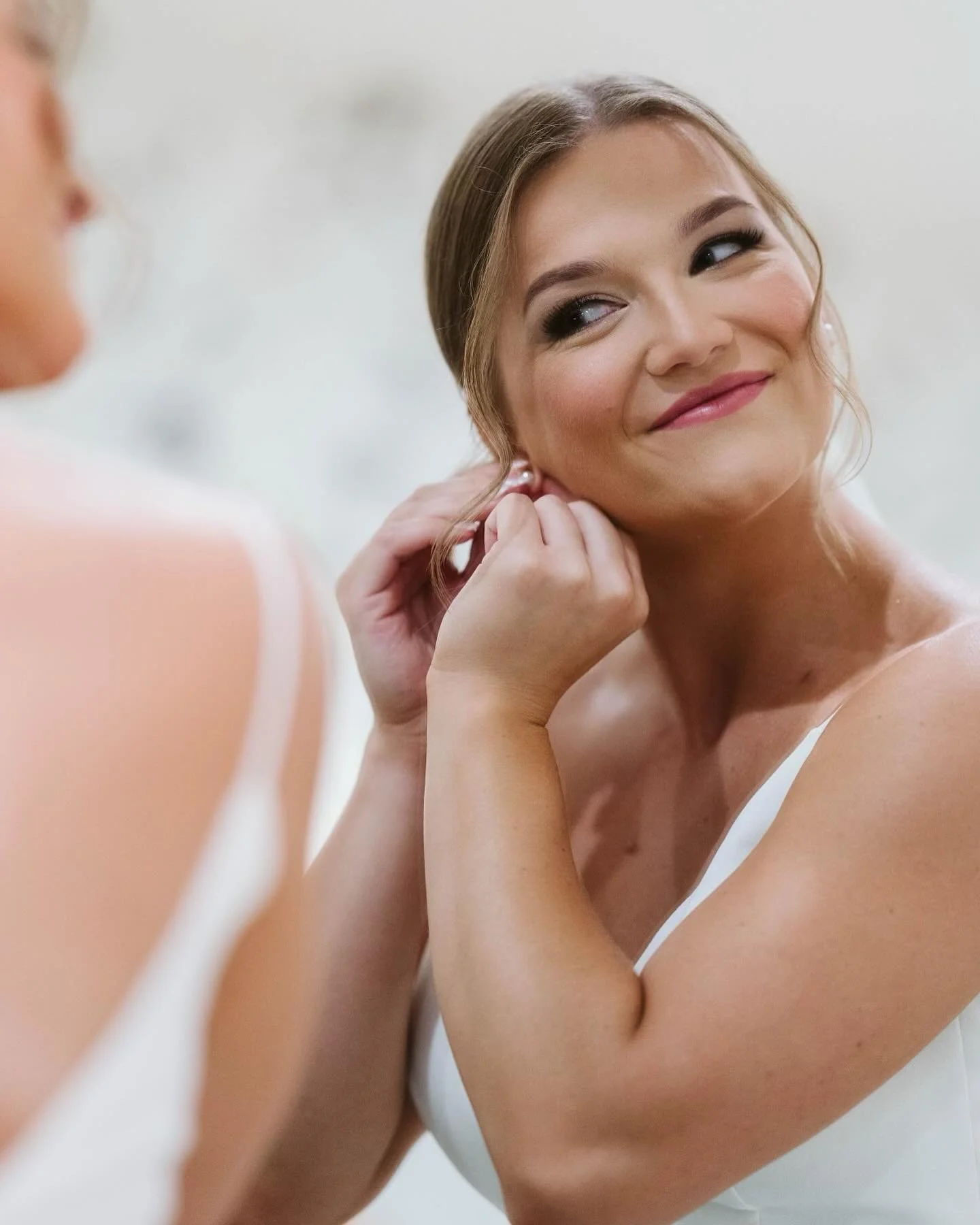 A woman with styled hair, makeup, and earrings, adjusting her earring in front of a mirror, smiling softly.
