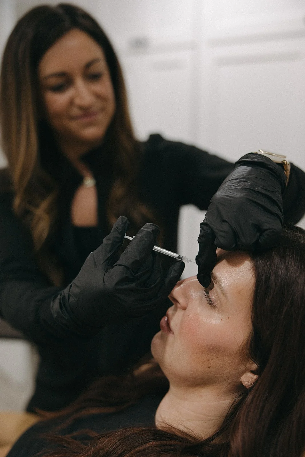 A woman receiving a cosmetic treatment with a syringe from a practitioner wearing black gloves.