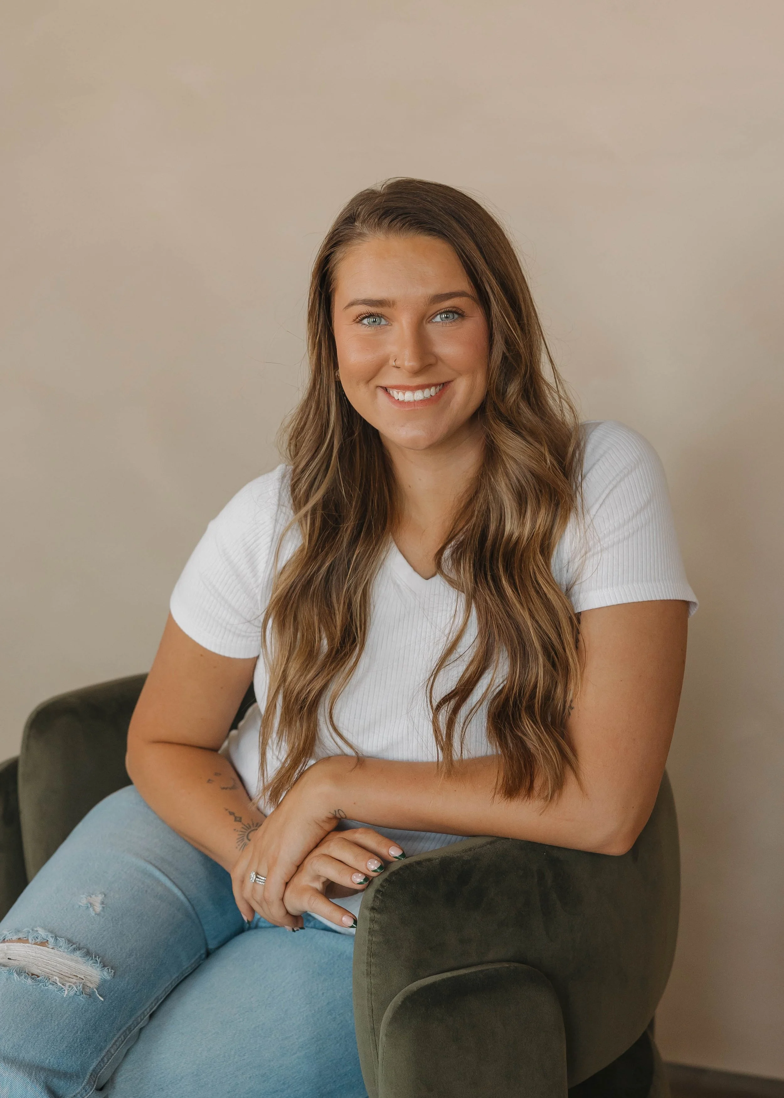 A woman with long wavy brown hair, blue eyes, and a nose piercing, smiling while sitting on a dark green armchair, wearing a white t-shirt and ripped blue jeans, against a neutral background.