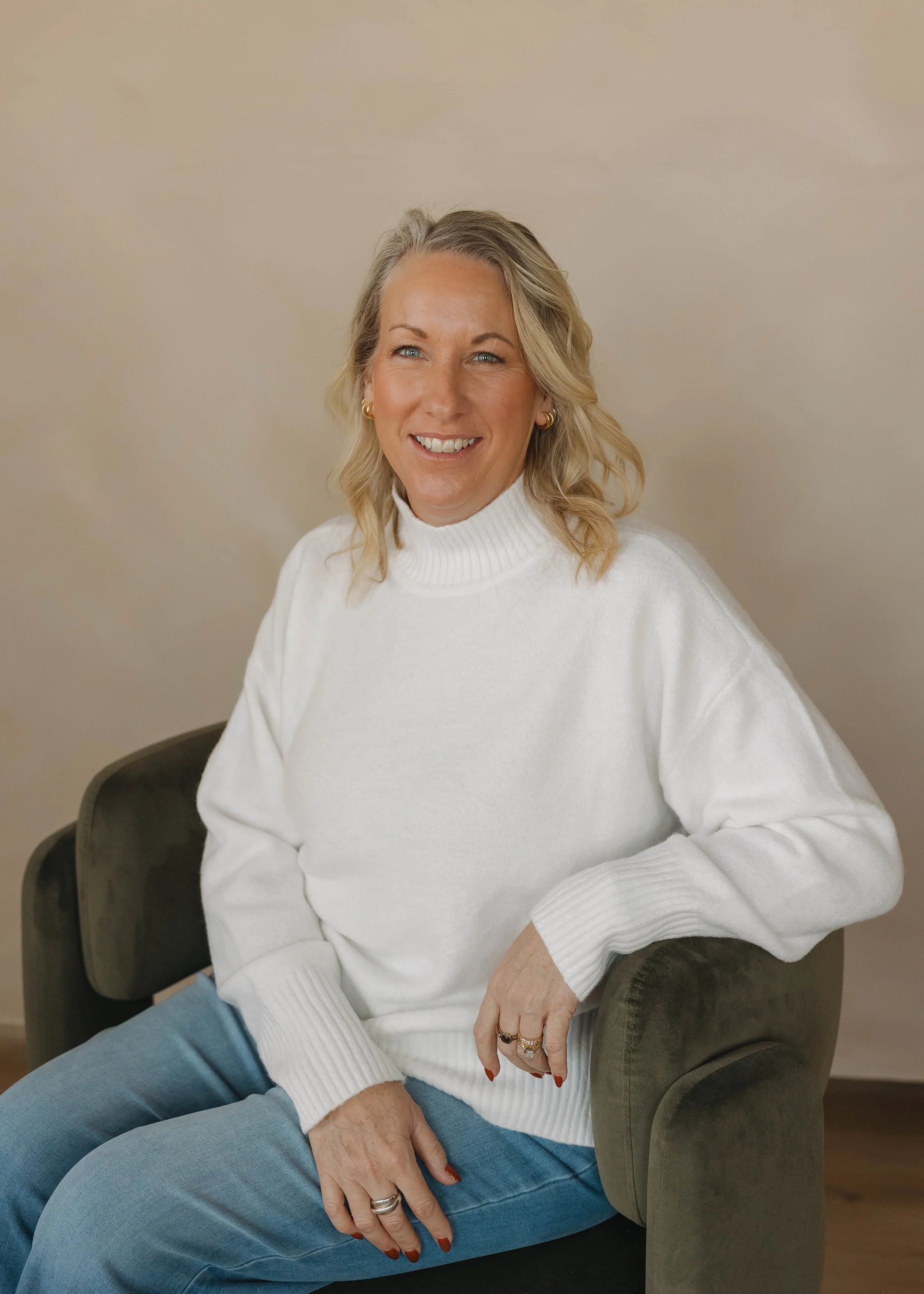A woman with blonde hair, wearing a white turtleneck sweater and blue jeans, sitting on a dark green chair and smiling at the camera.