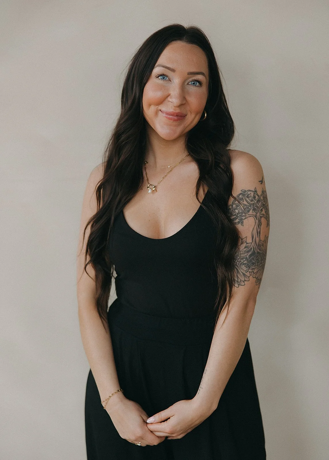 A woman with long dark hair, blue eyes, wearing a black sleeveless top, gold jewelry, and displaying a floral tattoo on her left arm, standing against a plain light-colored background.