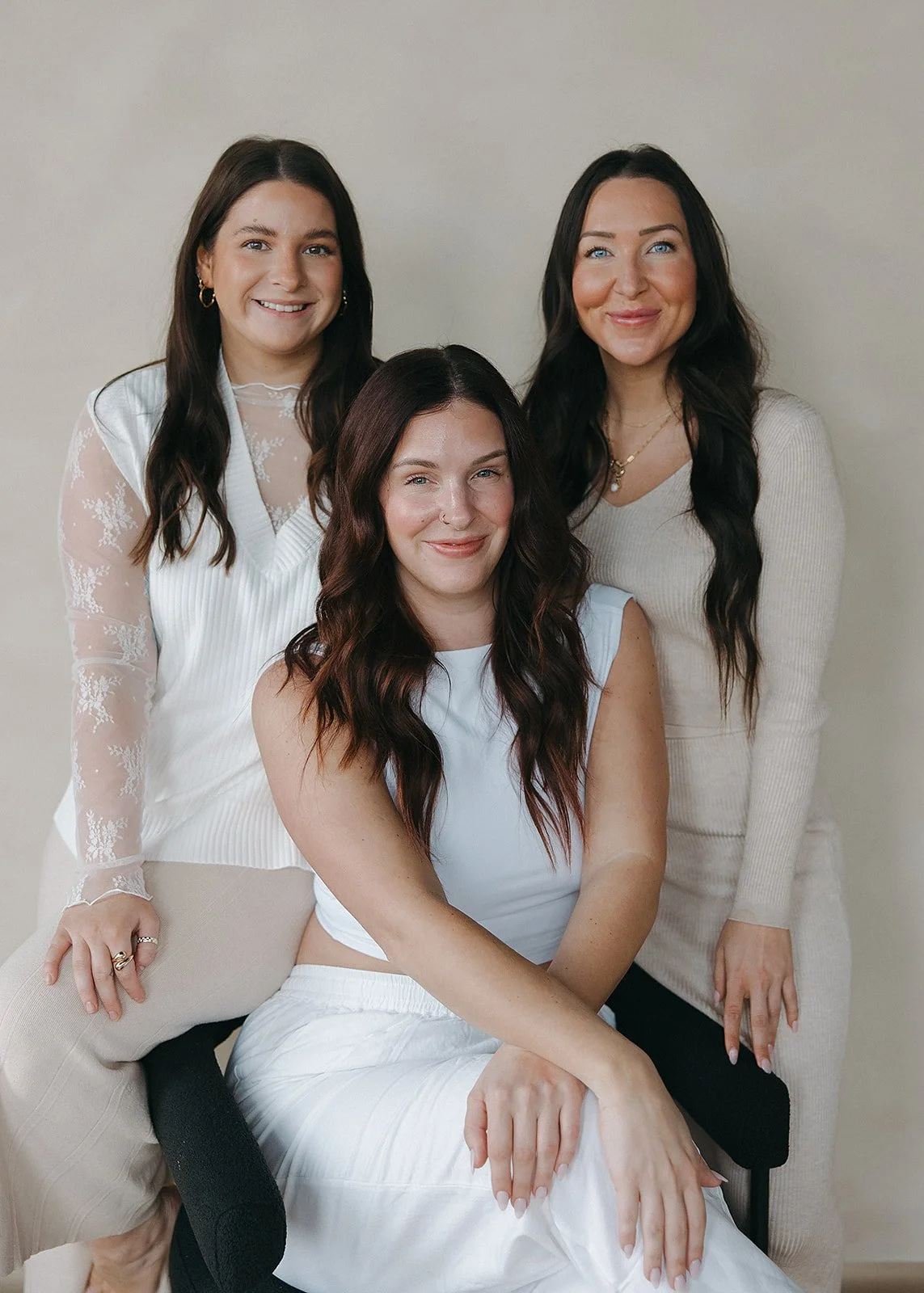 Four women with dark hair and light skin, dressed in white or cream, smiling at the camera against a plain beige background.