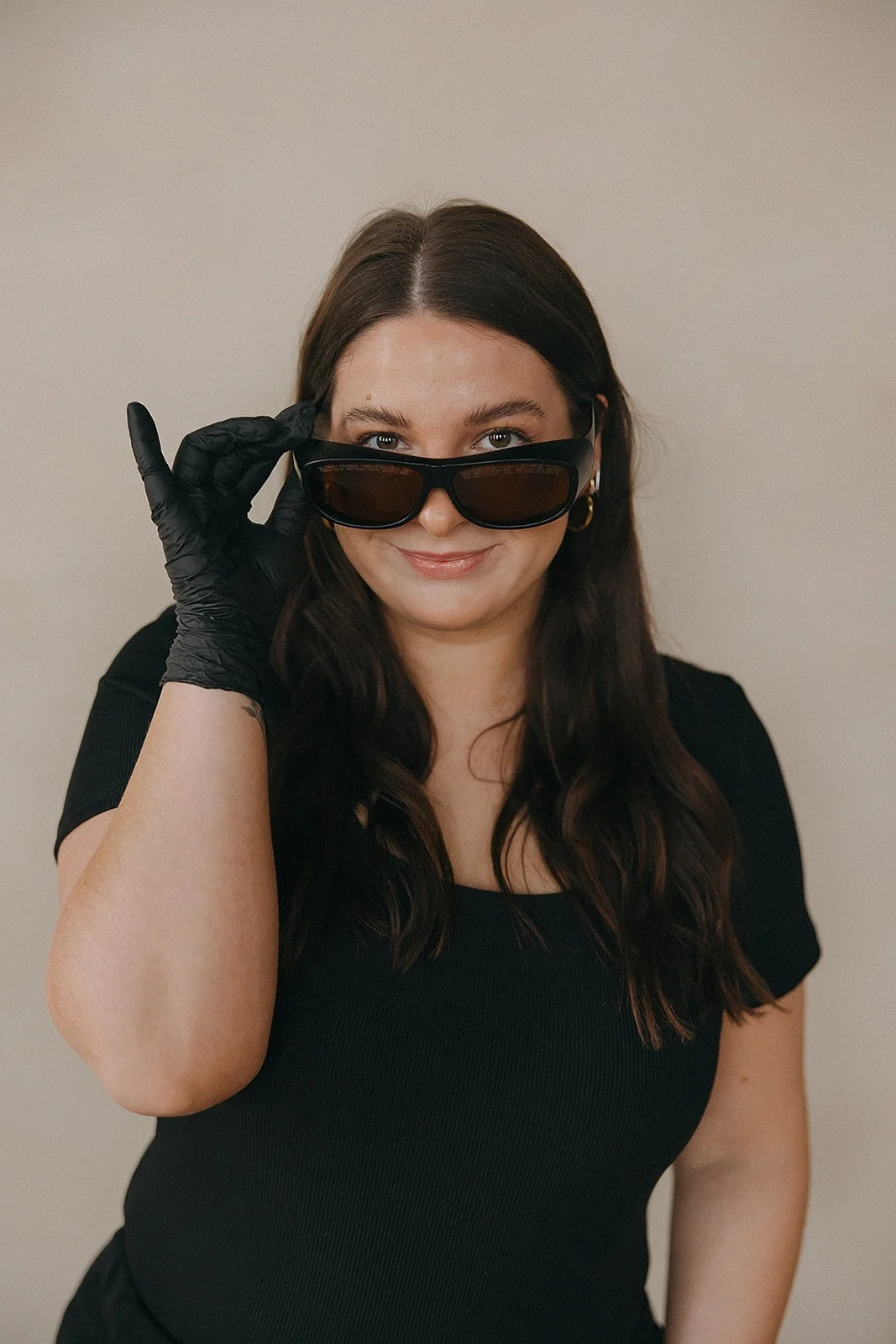 Woman with long dark hair wearing black shirt and black gloves, lifting sunglasses and smiling at camera.