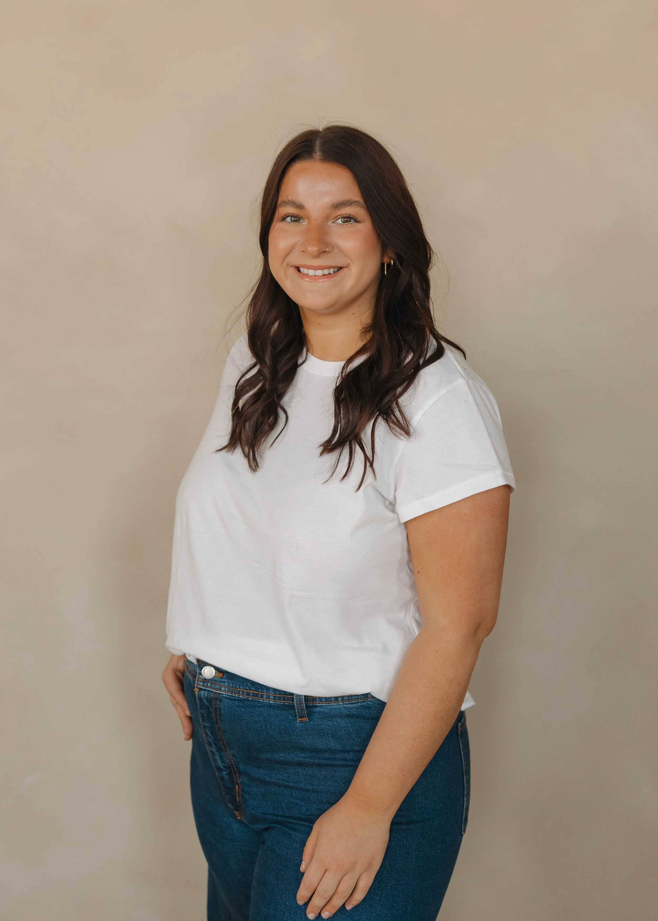 A woman with dark brown wavy hair, wearing a white t-shirt and jeans, smiling at the camera against a plain beige background.