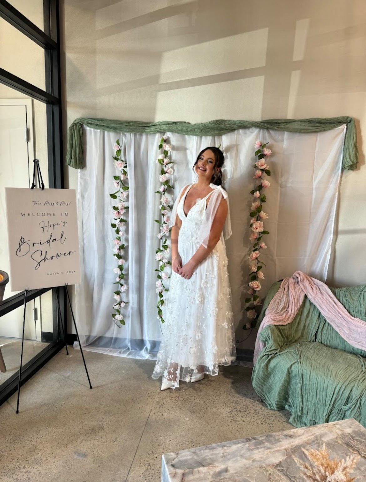 Bride in lace wedding dress standing in front of floral backdrop with sign for bridal shower.