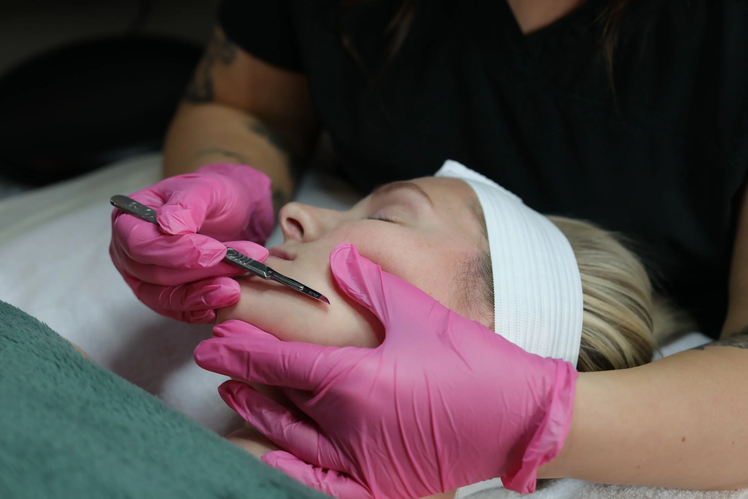 A woman with a headband lying on her back while a professional in pink gloves gives her a facial treatment with a small metallic tool.