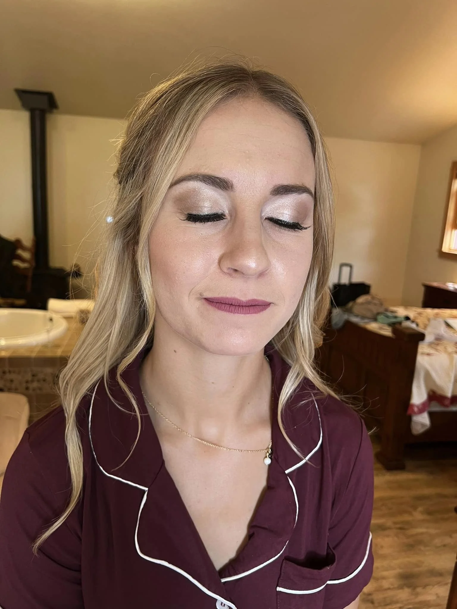A woman with blonde hair and closed eyes, wearing maroon pajamas with white piping and a delicate necklace, standing in a cozy bedroom.