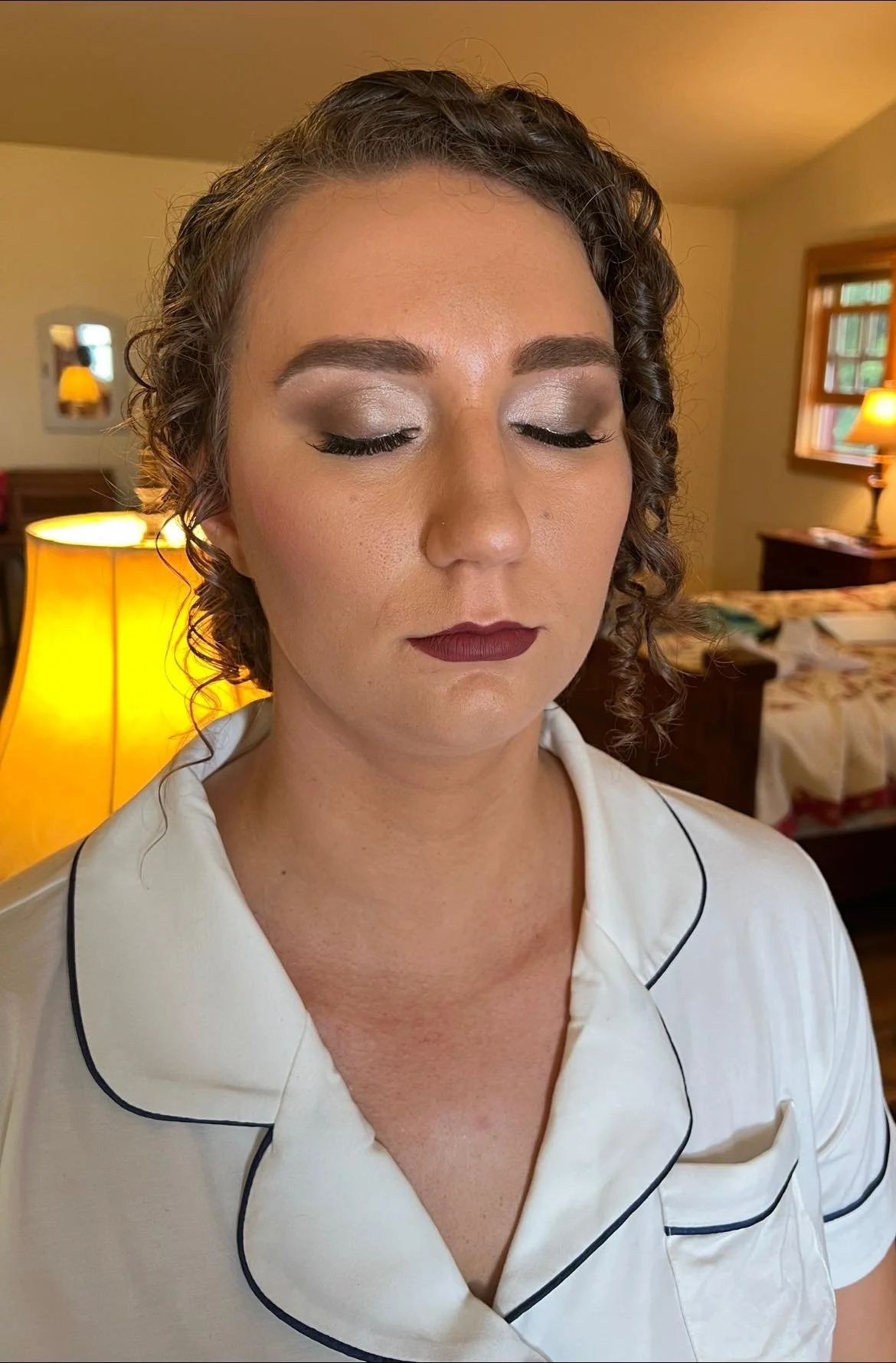 A woman with curly hair and makeup, wearing a satin pajama top with black piping, standing in a warmly lit bedroom with lamps and windows.