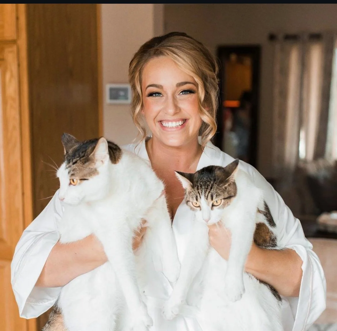 A woman with blonde hair smiling and holding two cats, one in each arm, indoors in a cozy room.