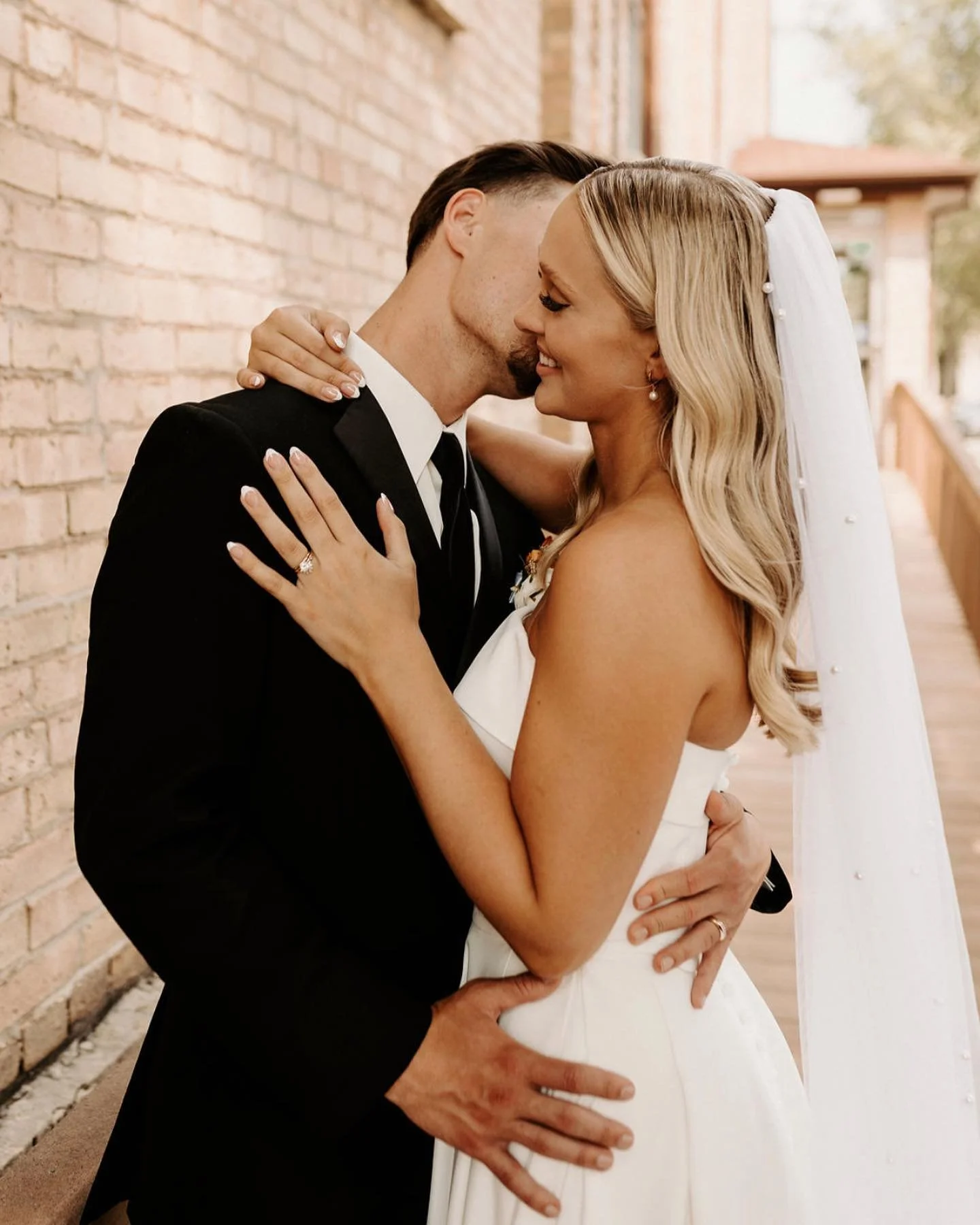 A newlywed couple sharing an intimate moment, embracing and kissing, outdoors on a wooden walkway beside a brick wall.