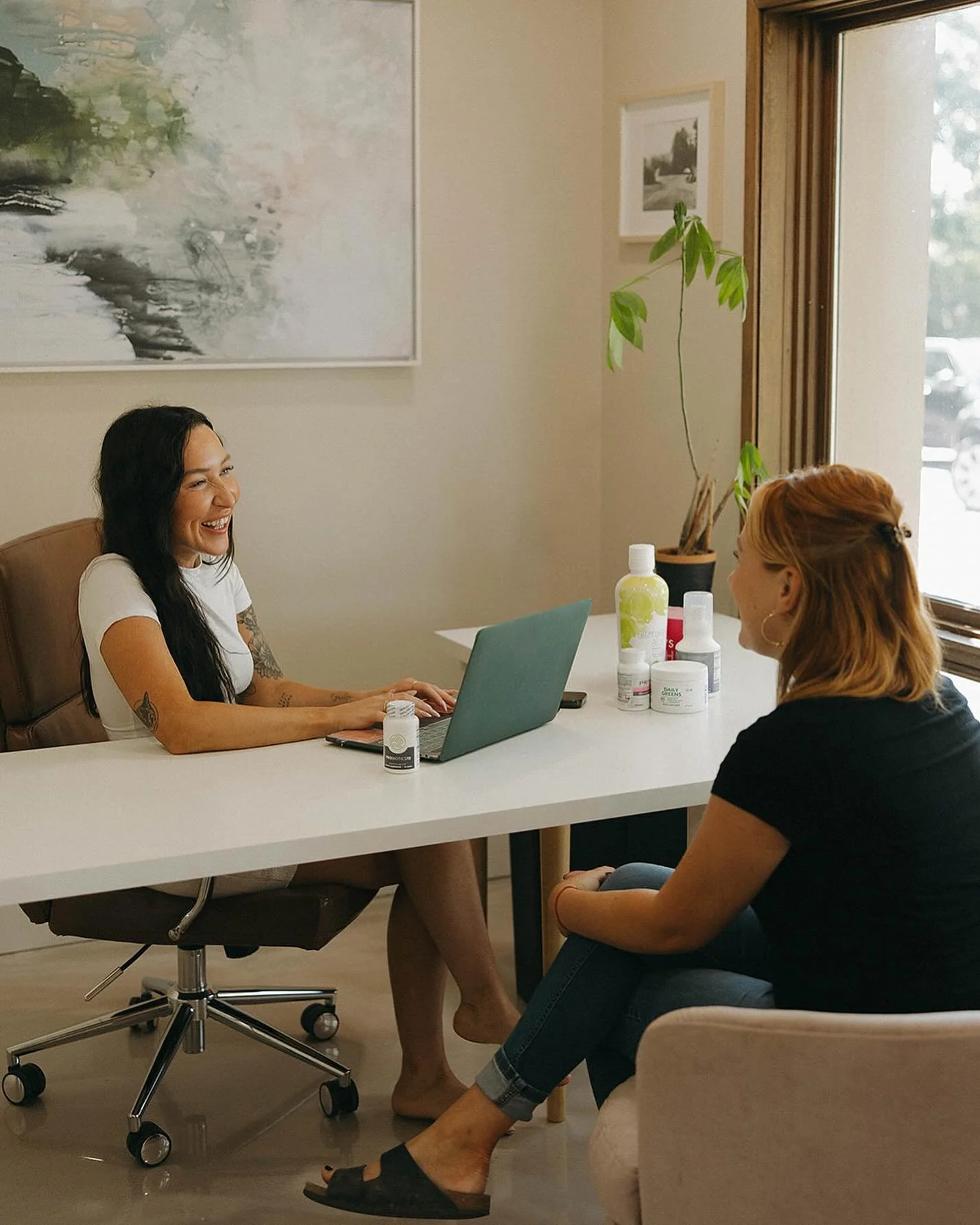 Two women having a friendly conversation in an office. One woman is sitting behind a desk with a laptop, smiling, and the other woman is seated in front of them. The desk has bottles and skincare products. A large window and a plant are in the background.