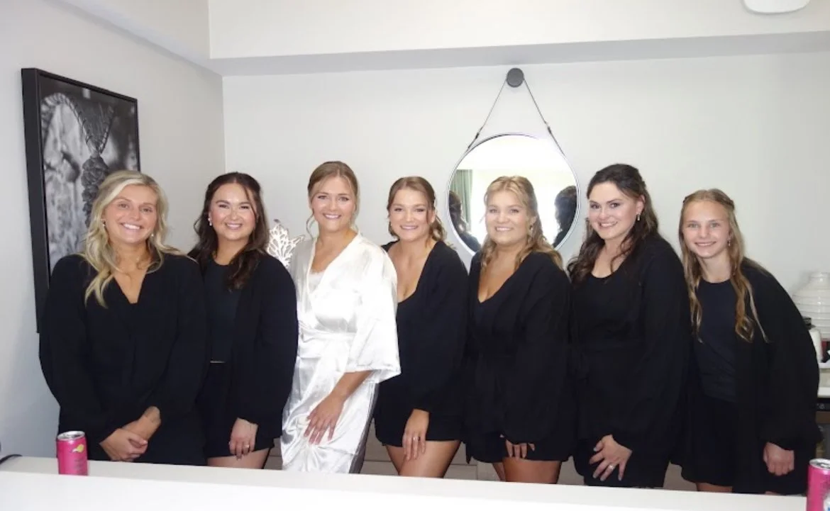 A group of seven women, mostly in black robes, standing together in a bedroom, smiling, with a woman in the center wearing a white robe, and a round mirror on the wall behind them.