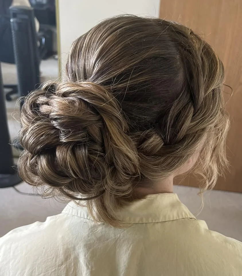 A woman with styled, braided and curled hair in an updo, wearing a light yellow shirt, in a room with a wooden door and some office furniture visible in the background.